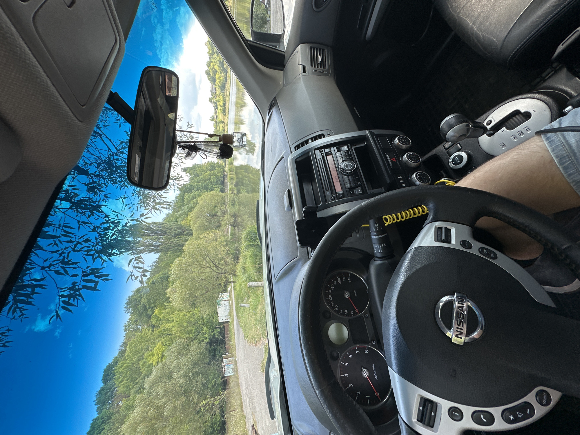 This is a first-person perspective inside a Nissan X-Trail, likely parked near a body of water in Makiv, Ukraine. The interior is dimly lit by sunlight filtering through the windshield. The dashboard features a steering wheel with Nissan branding and various controls, a central console with a radio and climate controls, and a gear shifter. A smartphone is mounted on the dashboard. Outside the vehicle, a calm lake or river is visible, surrounded by lush green trees and hills. The sky is a clear blue with scattered clouds. The overall impression is one of a peaceful, scenic stop during a road trip.