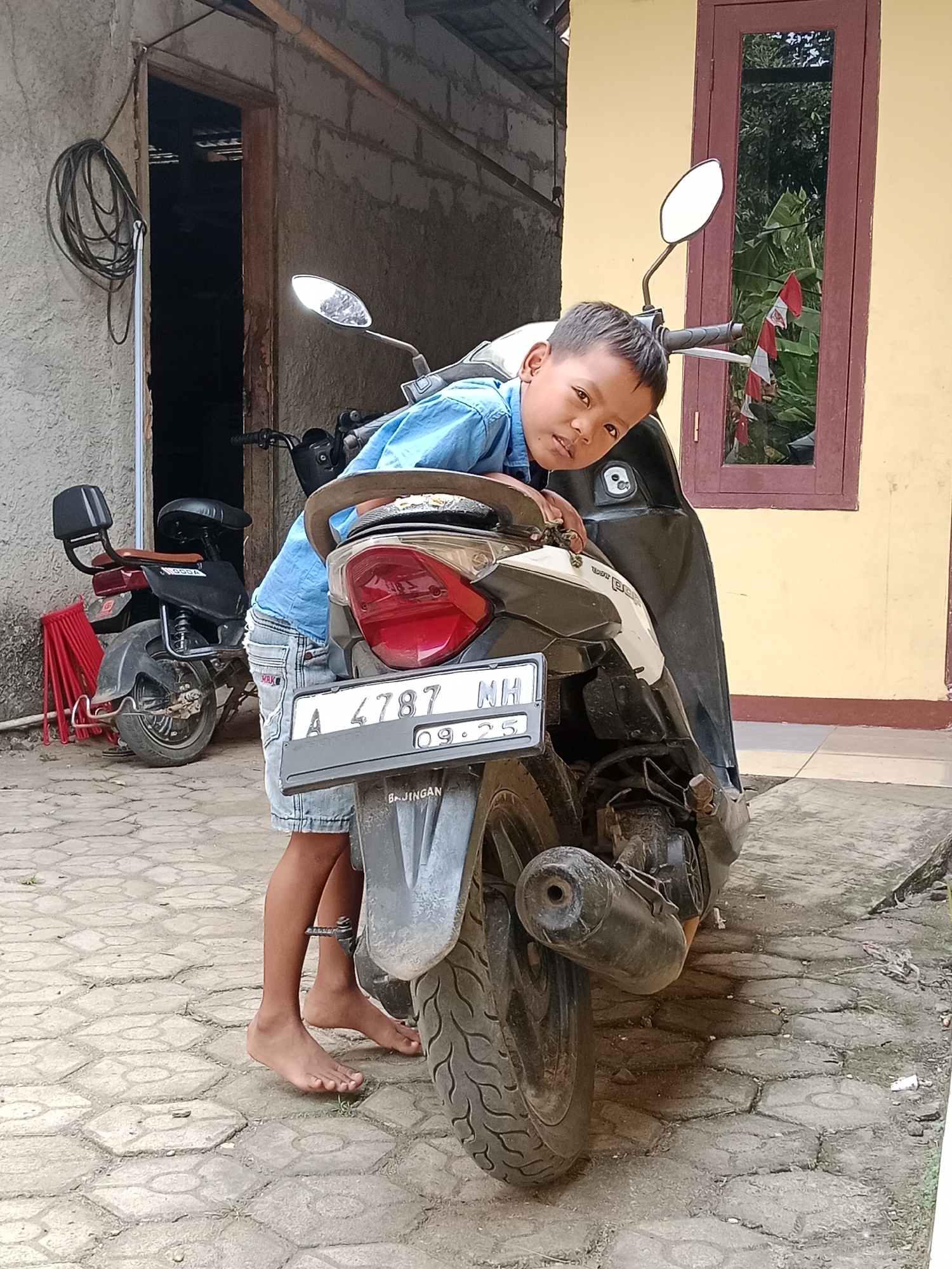 The image shows a young boy in Parage, Indonesia, leaning on a parked scooter. He is wearing a blue shirt and denim shorts, and is barefoot. The scooter is black and white, with a license plate that reads "A 4787 NH 09-25". In the background, there is a yellow building with a red-framed window, and a gray wall with a coiled black cable hanging on it. Another scooter, red and black, is parked to the left of the main subject. The ground is paved with interlocking hexagonal stones. The overall scene depicts a typical outdoor residential setting.