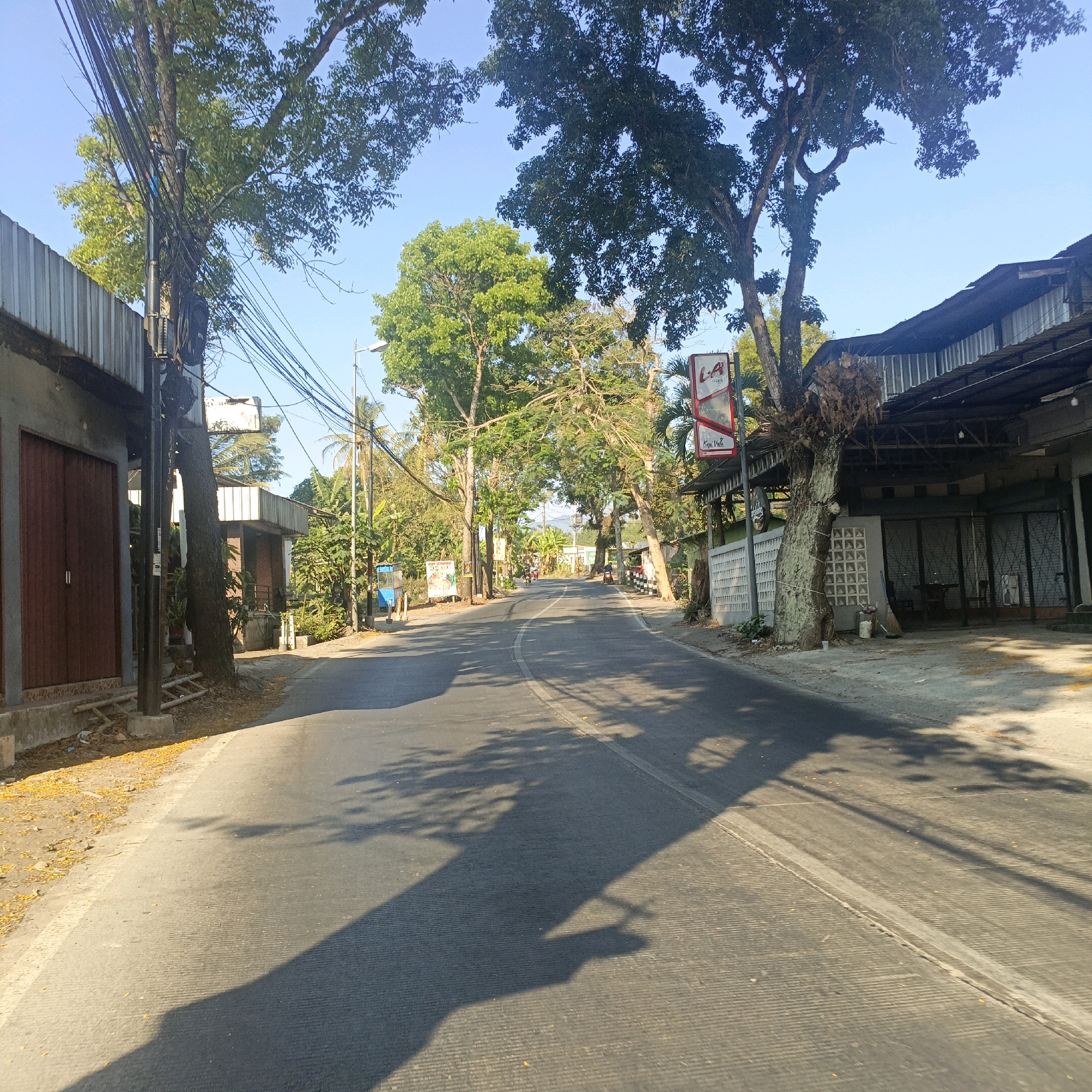 The image depicts a street scene in Salatiga, Indonesia. The road is paved and curves gently towards the distance, flanked by buildings and lush green trees. On the left side of the road, there is a building with a corrugated metal roof and large wooden doors. A utility pole covered in wires stands near this building. The right side of the road features a building with a similar roof and a white patterned wall. A red sign with "L.A Lights" and "Kopi Vale" is visible, attached to a tree. Shadows of the trees are cast across the road. Several motorcycles are visible in the distance, indicating some level of activity. The sky is clear and blue. The environment appears to be a semi-urban or suburban area with a tropical ambiance.