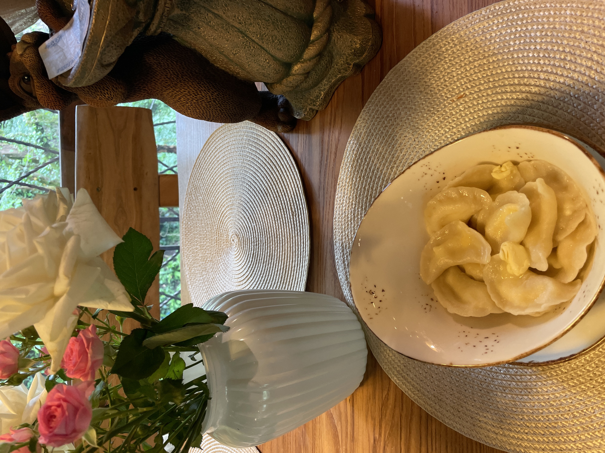 A top-down shot shows a wooden table setting. On the right side, a round, woven placemat supports a white plate filled with pierogi. Small dots are visible on the plate's rim. A pat of butter melts on top of the pierogi. To the left, another woven placemat sits beneath a white vase filled with white and pink roses. Behind the vase, a window is visible, showing glimpses of greenery. At the top left, a wooden statue rests on the table. The scene's location in Solokh-Aul, Russia, is only specified in the prompt and cannot be verified visually.