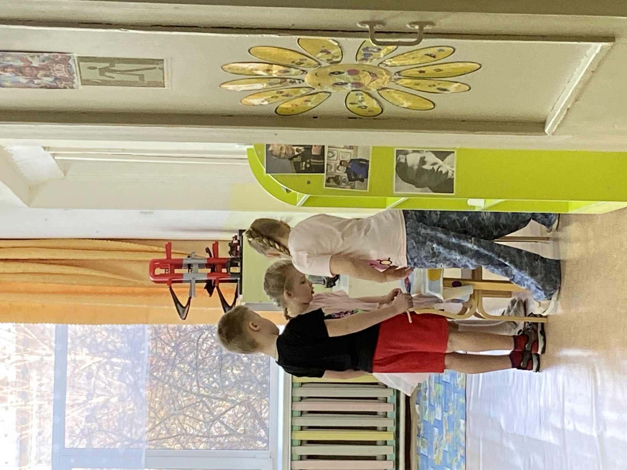 The image depicts three children standing in a room. A boy in a black shirt and red shorts stands in the front, with a girl in a pink shirt behind him. The girl in the back is wearing a white shirt and blue patterned pants. They appear to be engaged in some activity, possibly related to the wooden stick each of them is holding. The room has a window with a view of trees outside, a radiator, and colorful educational decorations. There is a decorative floral ceiling fixture. The location of this image is Noviy, Russia.