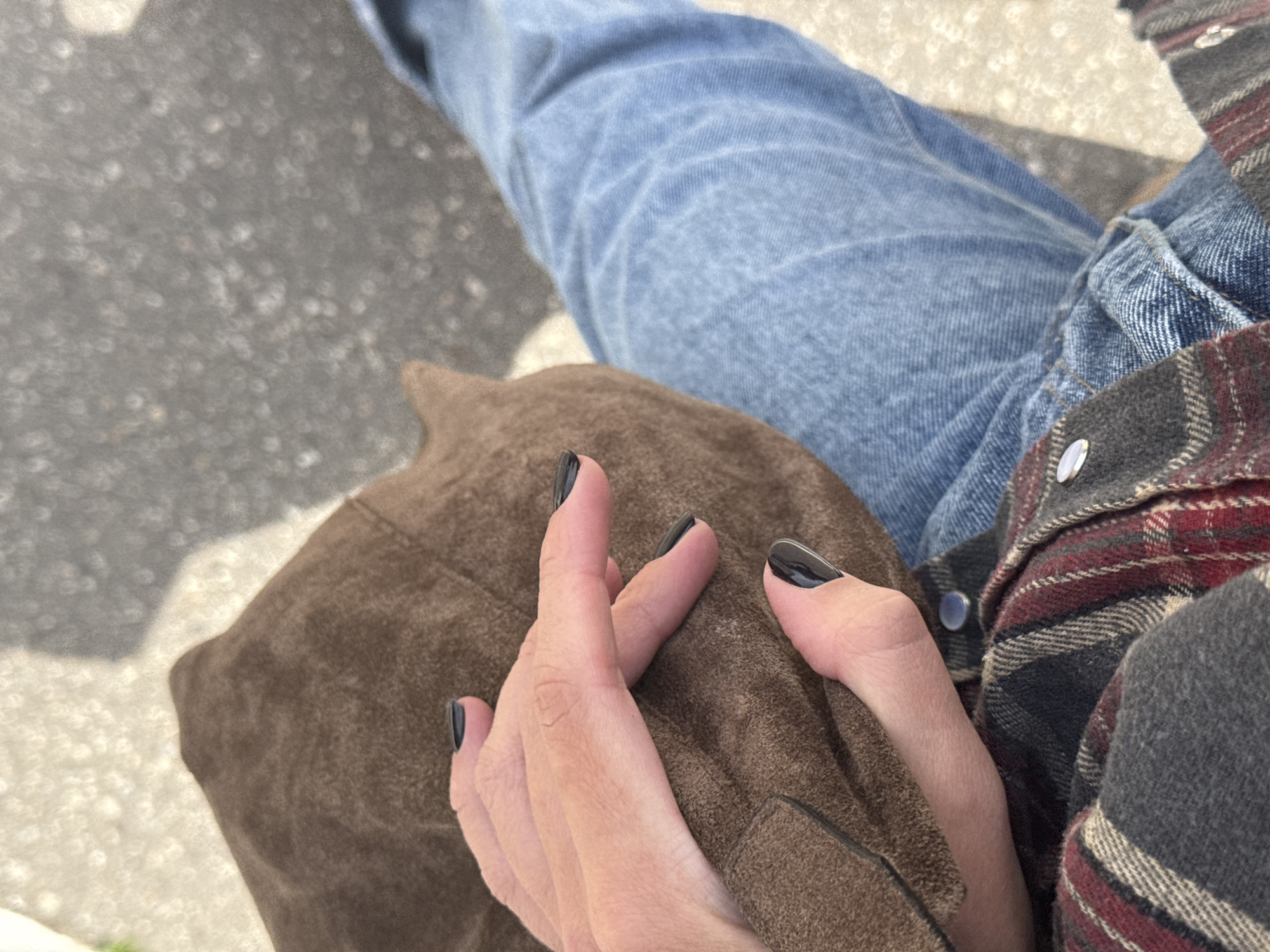 An overhead, close-up shot captures a moment between two individuals in Chaikovsky, Russia, likely outdoors during daylight hours. The scene focuses on the lower torso and legs of one person and the upper leg and hands of another, suggesting they are seated in close proximity.
One individual wears light blue denim jeans, visible on the right and top left of the frame. Their plaid flannel shirt, with shades of grey, red, and off-white stripes, and silver snap buttons, is visible on the right. Resting on the lap or between the two individuals is a textured, brown, suede-like garment or accessory.
The hands of the second person are prominent in the foreground, adorned with dark, possibly black, nail polish. Their fingers are subtly intertwined or resting gently on the brown material, suggesting a moment of connection or comfort. The bright sunlight creates strong illumination and highlights on the subjects, while the blurred background shows a light-colored ground surface, possibly asphalt or concrete, with a prominent bokeh effect indicating an outdoor setting. No specific text is visible in the image.