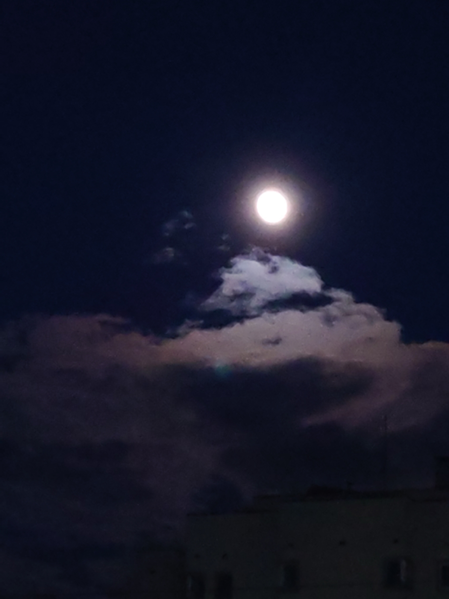 The image shows a full moon in a dark night sky. The moon is positioned near the top center of the frame and shines brightly. Partially obscuring the moon are dark, heavy clouds with lighter sections, indicating areas where the moonlight is able to penetrate. A building, with visible windows, is situated at the bottom of the frame. The location is in a city, though the specific city and location are unknown. There are no visible people in the scene.