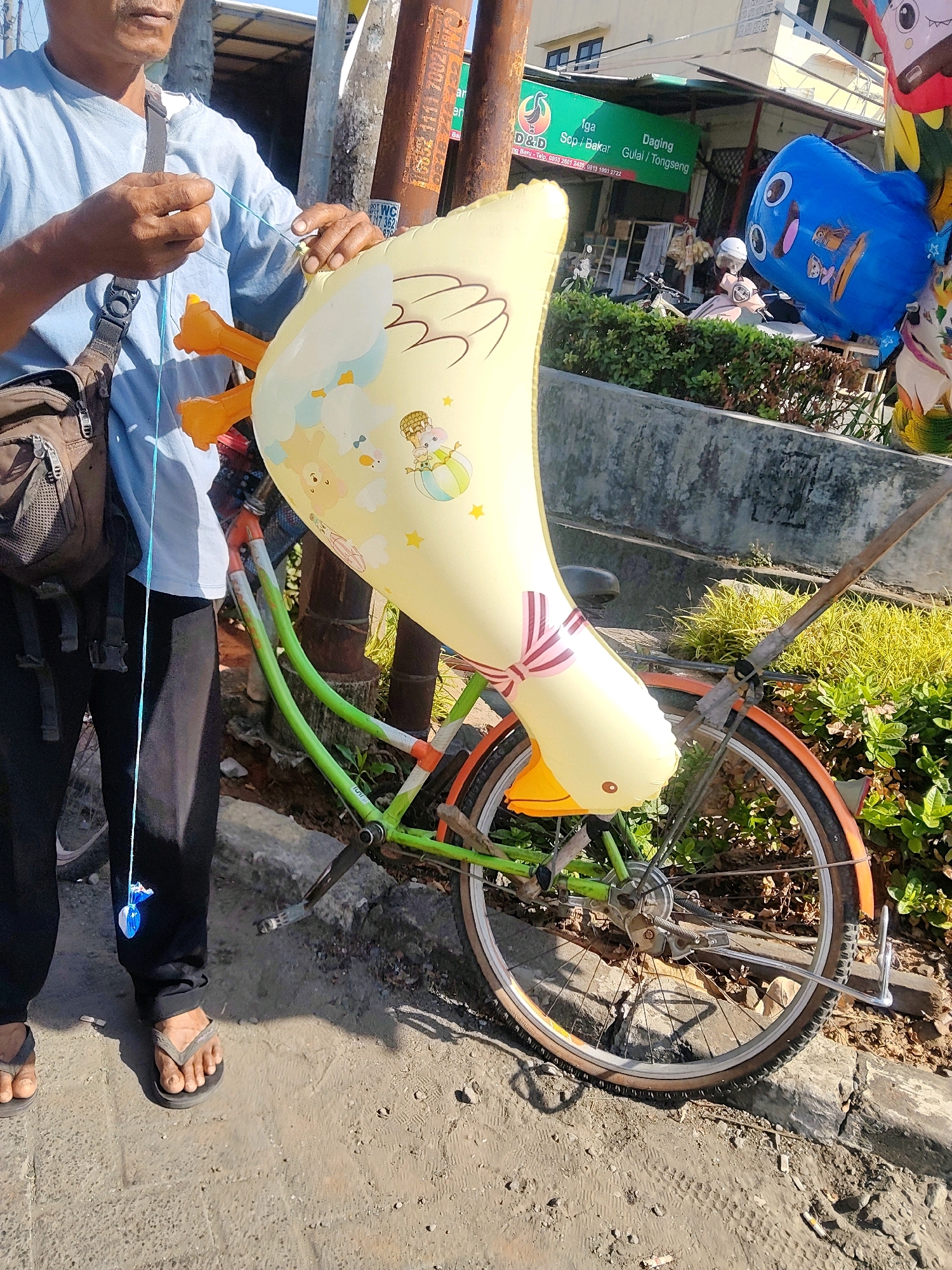 In Cibatu, Indonesia, a person stands next to a bicycle adorned with inflatable toys. The person, wearing a light blue shirt and black pants, is holding a blue string attached to a yellow inflatable toy shaped like a duck. The duck toy is decorated with cartoon characters and has orange feet. The bicycle has a green frame and orange accents on the rims and fender. In the background, a sign for a food establishment is visible, and several motorcycles are parked. The ground is paved, and there is lush greenery surrounding the scene.