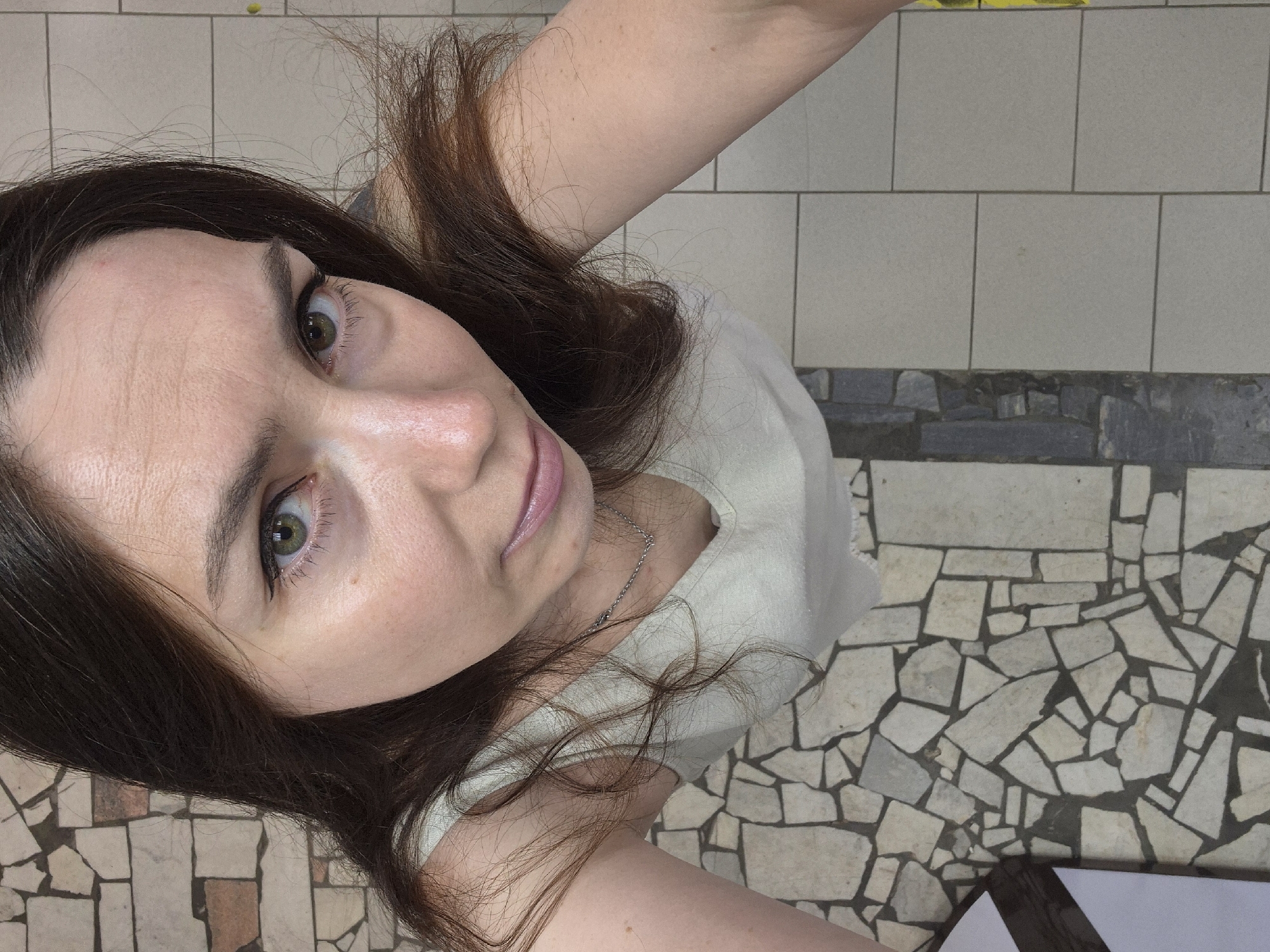 A high-angle, close-up shot captures a young adult female with long, dark brown hair. She is wearing a light-colored tank top and a thin silver chain necklace. Her gaze is directed upwards and slightly away from the camera. The background is composed of textured tiling, with a mosaic pattern of irregular stone pieces and a section of square tiles.