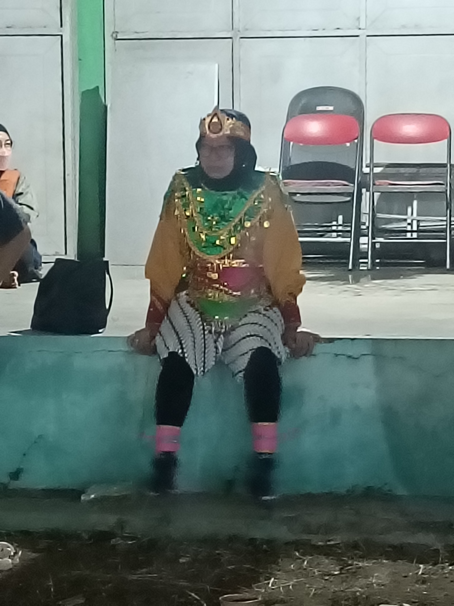 In the city of Kediri, Indonesia, an individual in traditional Javanese attire, adorned with a golden headdress and a green, gold, and red beaded vest, sits on a teal-colored concrete ledge. The individual wears a black hijab, black leggings, and striped pants with pink bands around the calves. In the background, a black backpack rests on the ledge to the left, and two red-seated chairs are visible on the right. The setting appears to be an outdoor area, possibly a performance space, with a plain wall and floor.