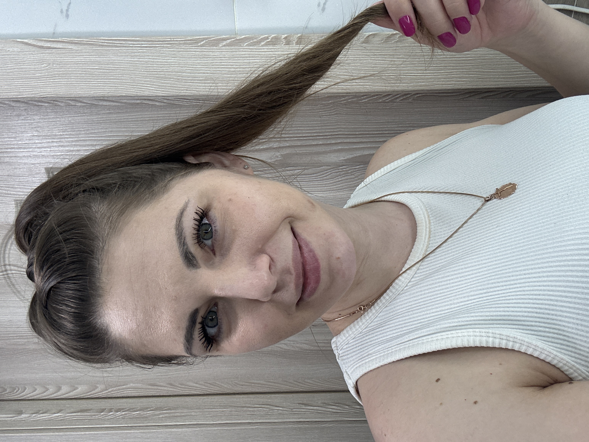 A young woman is depicted from the chest up, lying down and looking directly at the camera with a soft smile. Her head is positioned towards the bottom-left of the frame, indicating the image is rotated. She has long, brown hair, and playfully holds a strand of it with her right hand. Her fingernails are painted a vibrant magenta color.
She is wearing a white ribbed tank top and a thin gold chain necklace with a small, leaf-shaped pendant. Her eyes are light-colored, and she has prominent eyelashes. A small, sparkling stud earring is visible in her left ear. The background features light-colored wooden paneling or flooring, suggesting an indoor setting. The lighting is bright and even, consistent with daytime. Several small moles are visible on her right arm and shoulder area. There is no visible text in the image. The scene takes place in Раздолье, Russia.