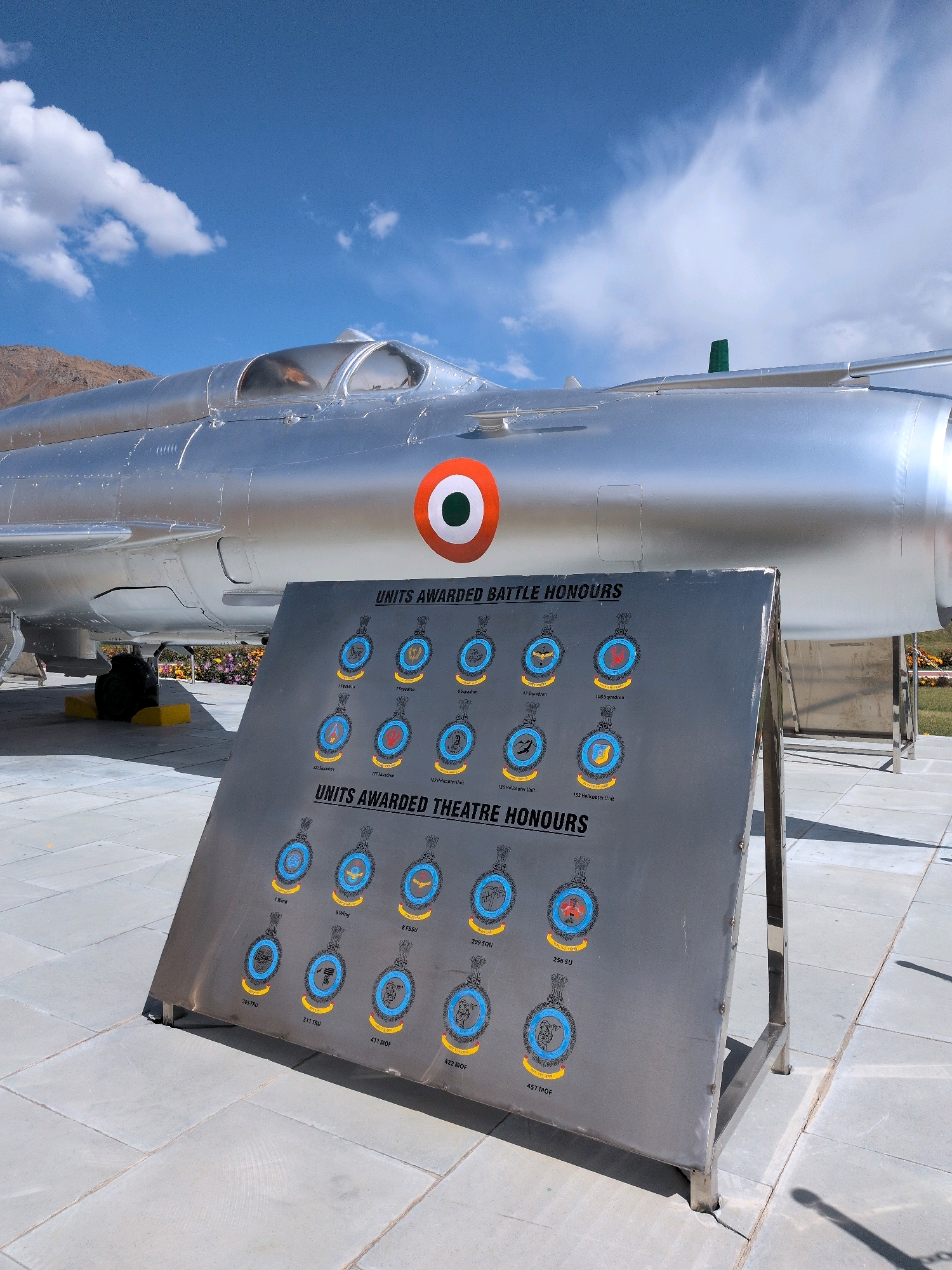 The image shows a silver fighter jet, likely a MiG-21, on display outdoors under a bright blue sky with scattered white clouds. The jet has an Indian Air Force roundel painted on its side. In front of the jet is a metal display board listing "UNITS AWARDED BATTLE HONOURS" and "UNITS AWARDED THEATRE HONOURS." The board features numerous circular emblems, each with a unit designation below it. The scene appears to be a memorial or museum in a public space, possibly related to military history. The ground is paved with large grey tiles. In the background, there are hints of mountains and some flowering bushes. The location is identified as Bhimbat, India.