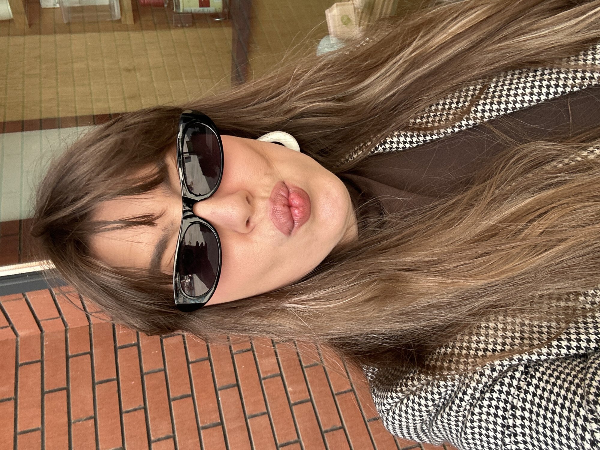 A close-up, rotated portrait captures a young woman against an urban backdrop, likely in Tbilisi, Georgia. Her long, wavy brown hair frames her face, flowing over her shoulders and jacket. She wears large black-framed sunglasses, obscuring her eyes, and a small, white circular earring is partially visible on her right ear. Her lips are slightly pursed in a relaxed expression.
She is dressed in a stylish black and white houndstooth patterned jacket, worn over a dark brown turtleneck or top, with the distinct pattern visible around her shoulder. The background features a prominent red brick wall in the lower left and bottom of the frame, suggesting an outdoor or street-level setting. Above and behind her, a lighter, more reflective surface with a grid-like pattern can be seen, possibly an indoor floor or a reflection through a window. The lighting indicates it is daytime. There is no visible text.