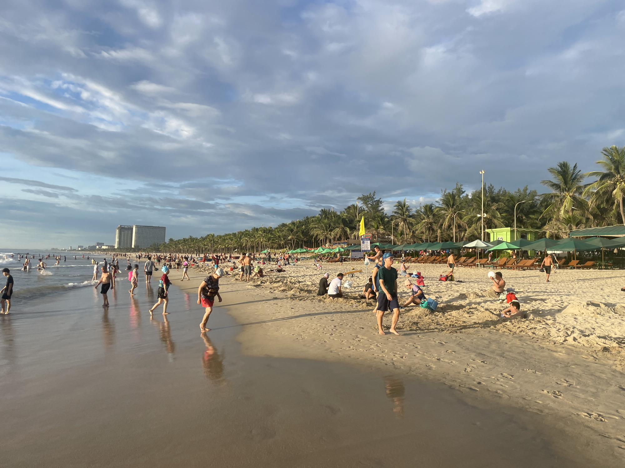 This is a photograph of a crowded beach, likely My Khe Beach in Da Nang, Vietnam, taken during the daytime under a partly cloudy sky. The foreground shows the wet sand of the shoreline, reflecting the sky and the people on the beach. The middle ground is populated by numerous beachgoers engaged in various activities. Some are wading in the shallow water, while others are walking on the sand. Several groups are gathered on the dry sand, with some individuals appearing to play or rest. Further back, a line of palm trees separates the beach from a resort area, where lounge chairs and umbrellas are set up. In the distance, a tall hotel building is visible, and the overall impression is one of a lively and popular tourist destination. The lighting suggests it could be late afternoon, with long shadows beginning to form.