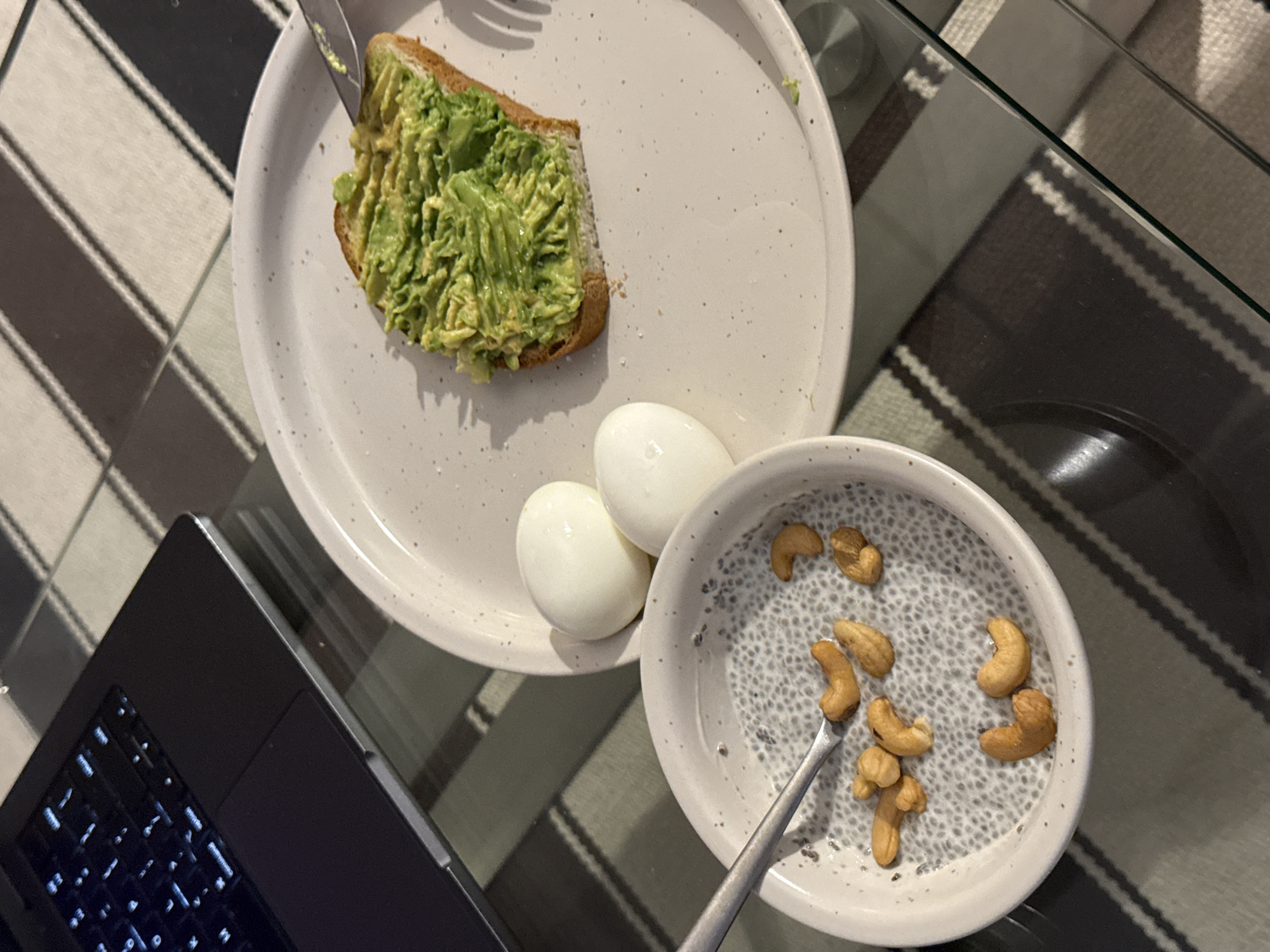 The image shows a meal consisting of avocado toast, two hard-boiled eggs, and a bowl of chia seed pudding topped with cashews, arranged on a speckled white plate and a smaller bowl. A fork rests on the plate near the toast, and a spoon is in the bowl. A laptop sits to the left, partially in frame. The meal is placed on a glass-topped table, which sits on a striped rug. The location is unknown.