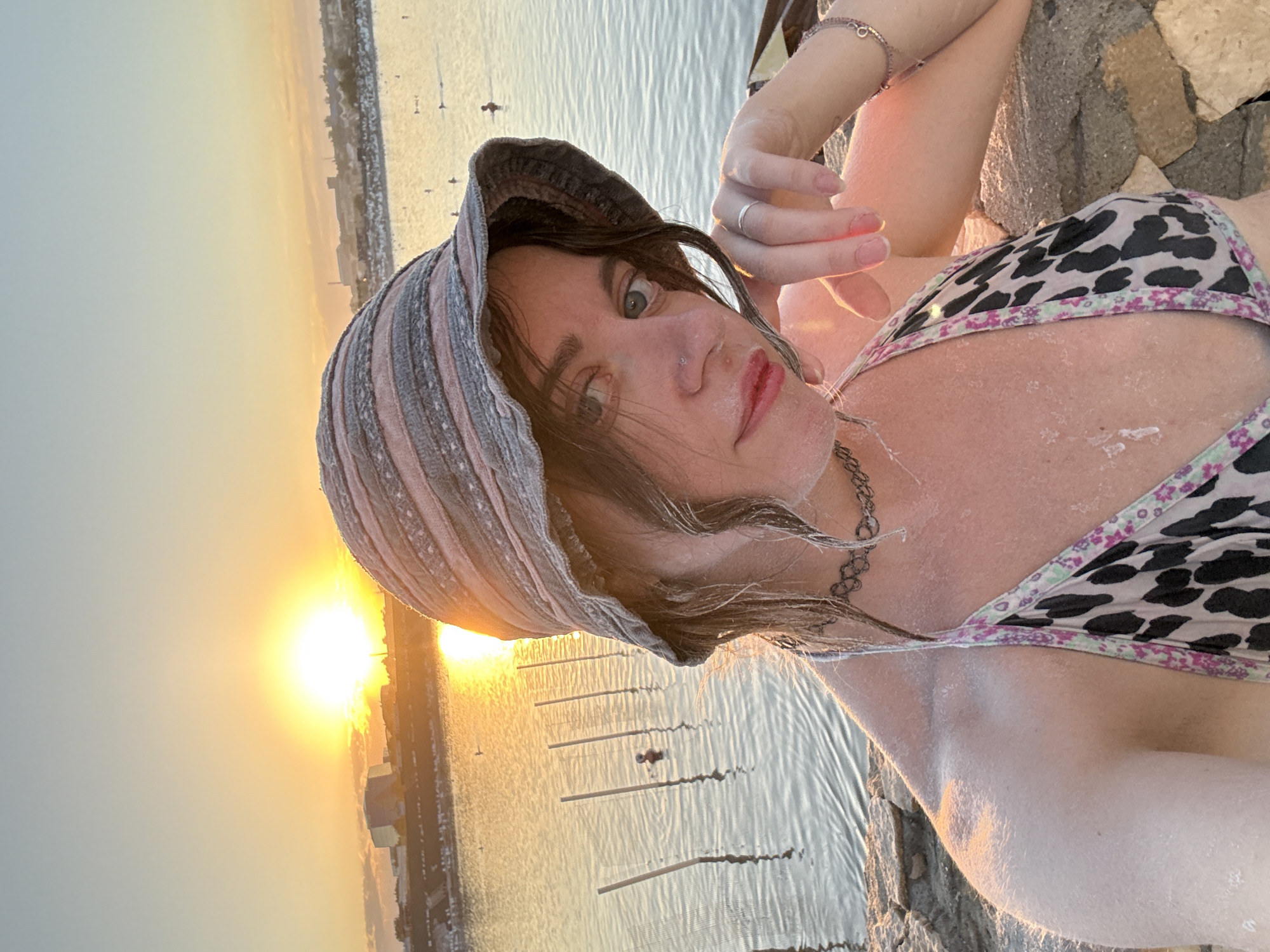 A woman is taking a selfie at sunset near a body of water. She is wearing a bucket hat and a bikini. Her skin is covered in a white substance, likely salt, as the city of Sol-Iletsk, Russia is known for its salt lakes and salt mining. In the background, the sun is setting over a city skyline, casting a warm glow on the water. There are several small figures in the water, suggesting other people are also enjoying the area. Wooden posts are visible in the water, possibly marking a designated swimming or fishing area. The woman is resting her chin on her hand and looking directly at the camera.