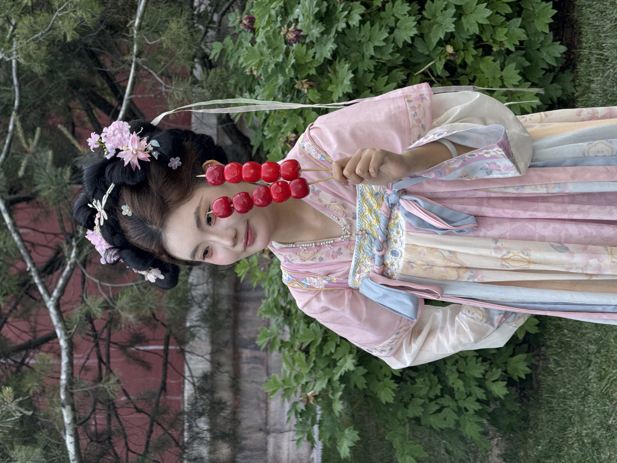 A young woman dressed in a traditional Chinese hanfu garment poses in an outdoor setting. She is holding two skewers of red, glazed fruit. The backdrop features a blurred landscape of greenery and glimpses of structures. The location is possibly Dongcheng District, China.