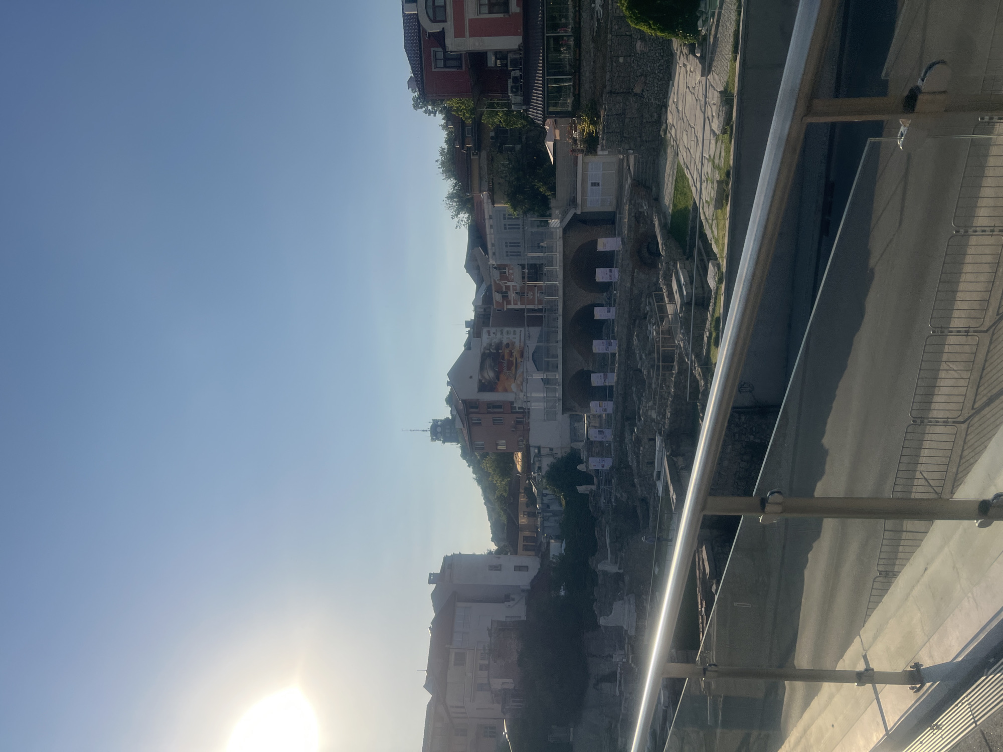 The image captures an outdoor view of the Roman Stadium of Philippopolis in Plovdiv, Bulgaria. The sun is high in the sky, casting long shadows and creating a bright, clear atmosphere. In the foreground, a modern metal railing and glass barrier are visible, suggesting a viewing platform or walkway. Beyond this, the ancient ruins of the Roman Stadium are laid out, with visible stone structures, seating areas, and remnants of arches. Several informational posters are placed along the stadium's perimeter.
Behind the stadium, the cityscape of Plovdiv unfolds. A cluster of buildings, a mix of older and more modern architecture, are situated on a gentle slope. The buildings display various colors, including white, pink, red, and earthy tones. Notably, a tall tower with antennae is visible on a hill in the distance, overlooking the city. Green trees are interspersed among the buildings, indicating a city park or natural vegetation. A single person, appearing as a small figure in the distance, is walking through the archaeological site. The overall scene depicts a historical site integrated into a contemporary urban environment.