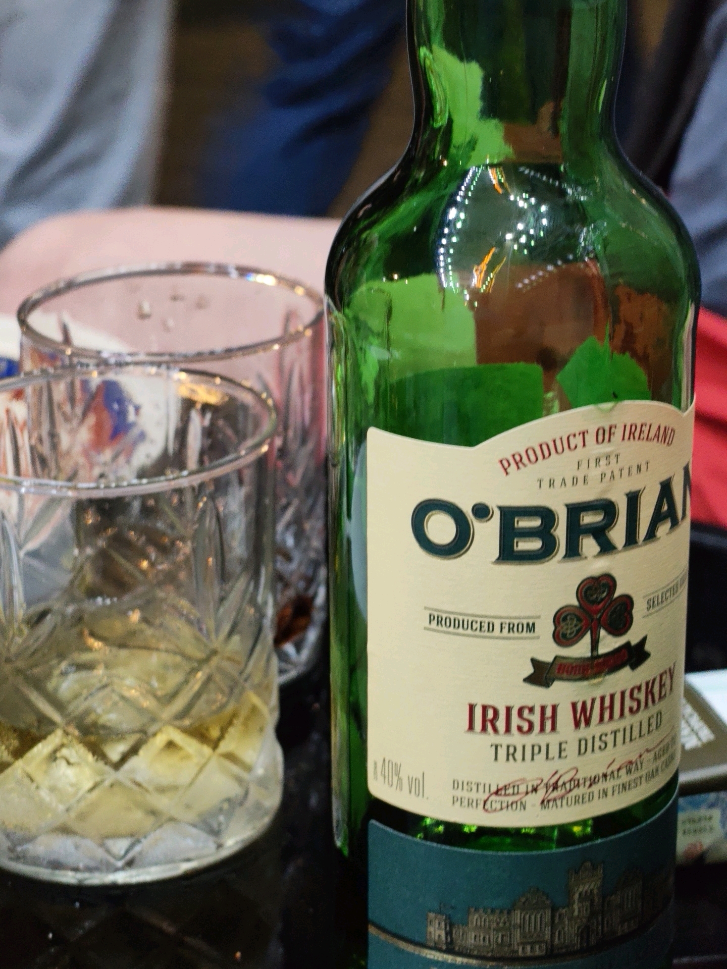 This image shows a close-up of a green bottle of O'Brian Irish Whiskey and two glasses of whiskey on a dark surface. One glass is in the foreground, filled with whiskey and ice, and the other is behind it, also containing whiskey. The bottle has a label that reads "PRODUCT OF IRELAND", "FIRST TRADE PATENT", "O'BRIAN", and "IRISH WHISKEY TRIPLE DISTILLED". The background is blurred and shows parts of people's clothing. The scene appears to be indoors, possibly in a bar or restaurant. There is no visible indication of the specific location within the city of Batumi, Georgia.