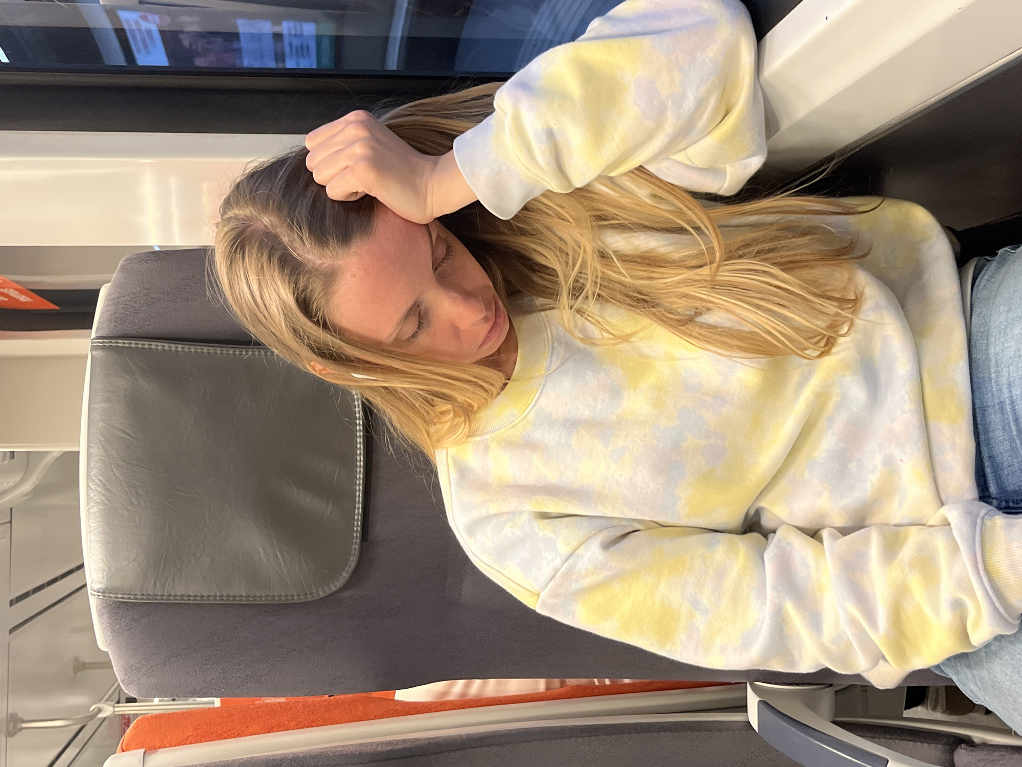 A woman is sleeping on a train. She is slumped against the window with her hand resting on her head. She is wearing a white and yellow tie-dye sweatshirt and blue jeans. The train car appears to be modern and clean. There is a window, a gray headrest, and an orange tray table behind her. The location cannot be confirmed as Barcelona, Spain.