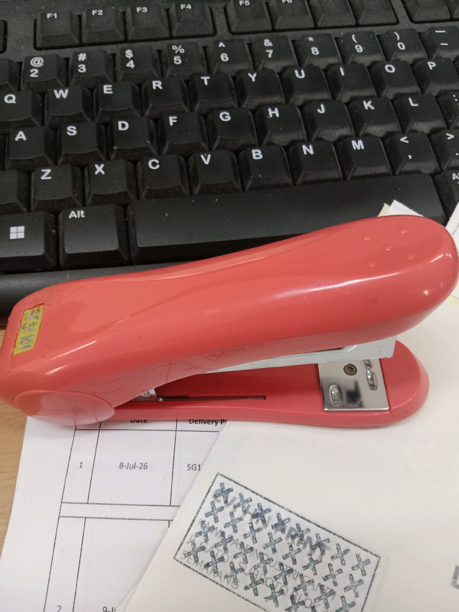 The image shows a pink stapler placed on a desk in front of a keyboard. A document with printed lines and some text is visible beneath the stapler, along with a stamped piece of paper featuring a pattern of "X" marks and the word "SEMARANG" which indicates the location is Semarang, Indonesia. The scene appears to be an office or workspace environment. There are no people visible in the image.