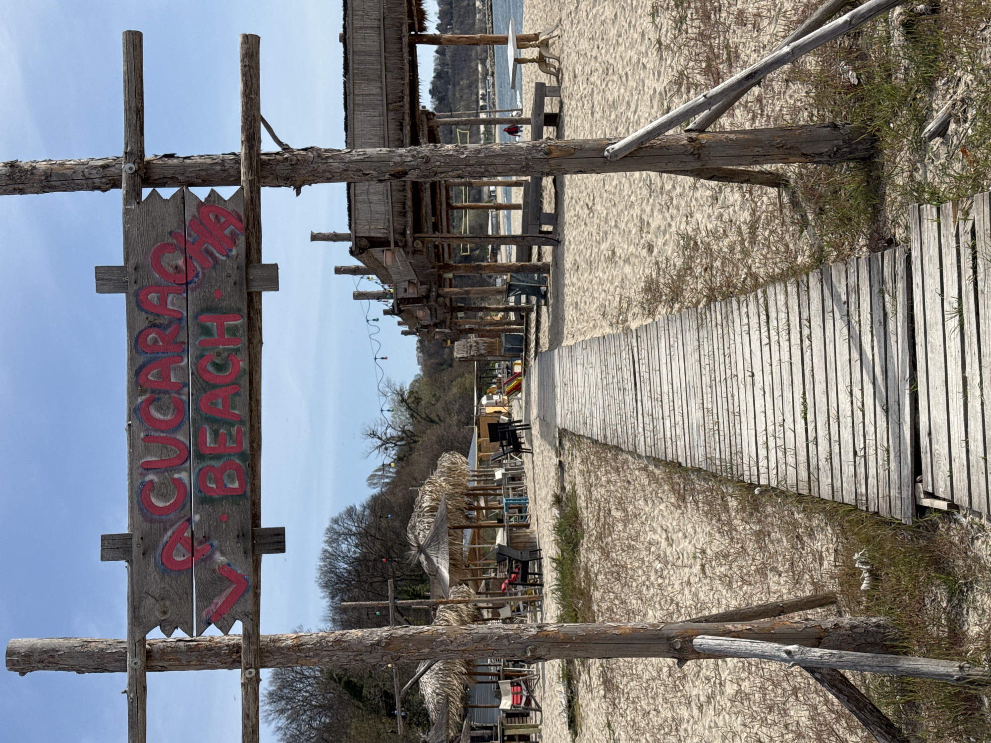 This image captures a beach scene in Varna, Bulgaria. A weathered wooden sign reading "V. Cucaracha Beach" stands prominently in the foreground. Behind the sign, a wooden boardwalk stretches towards the beach, flanked by sandy areas with sparse vegetation. Beachside structures with thatched roofs and wooden supports line the pathway. In the distance, a glimpse of the sea is visible under a blue sky with light cloud cover.