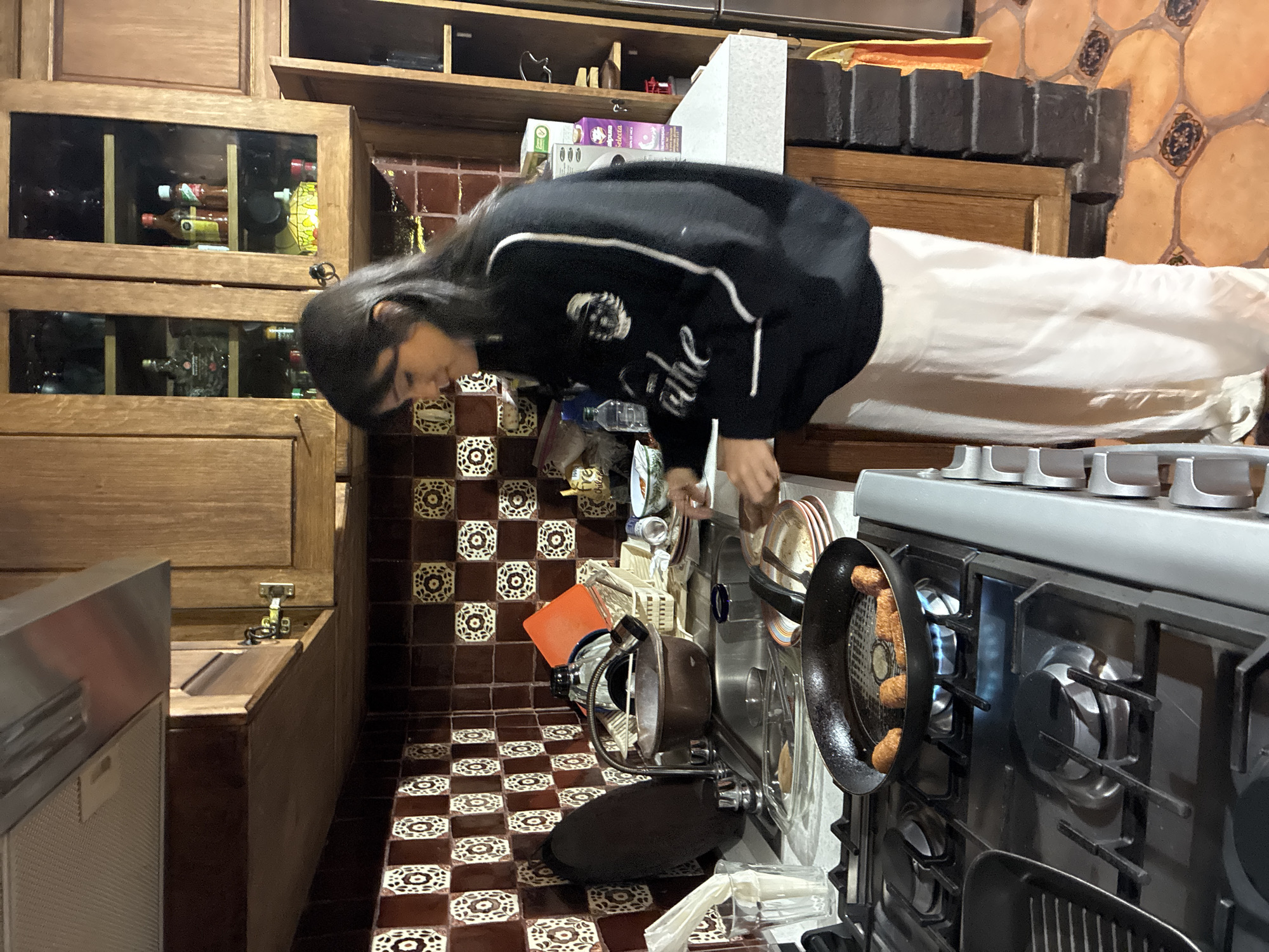 A person with long dark hair, wearing a black sweatshirt and white pants, is bending over a sink in a kitchen. The kitchen features brown tiled flooring and walls, and wooden cabinets. A stove with a frying pan containing food is to the right of the sink. The location is Cañada de Madero, Mexico.