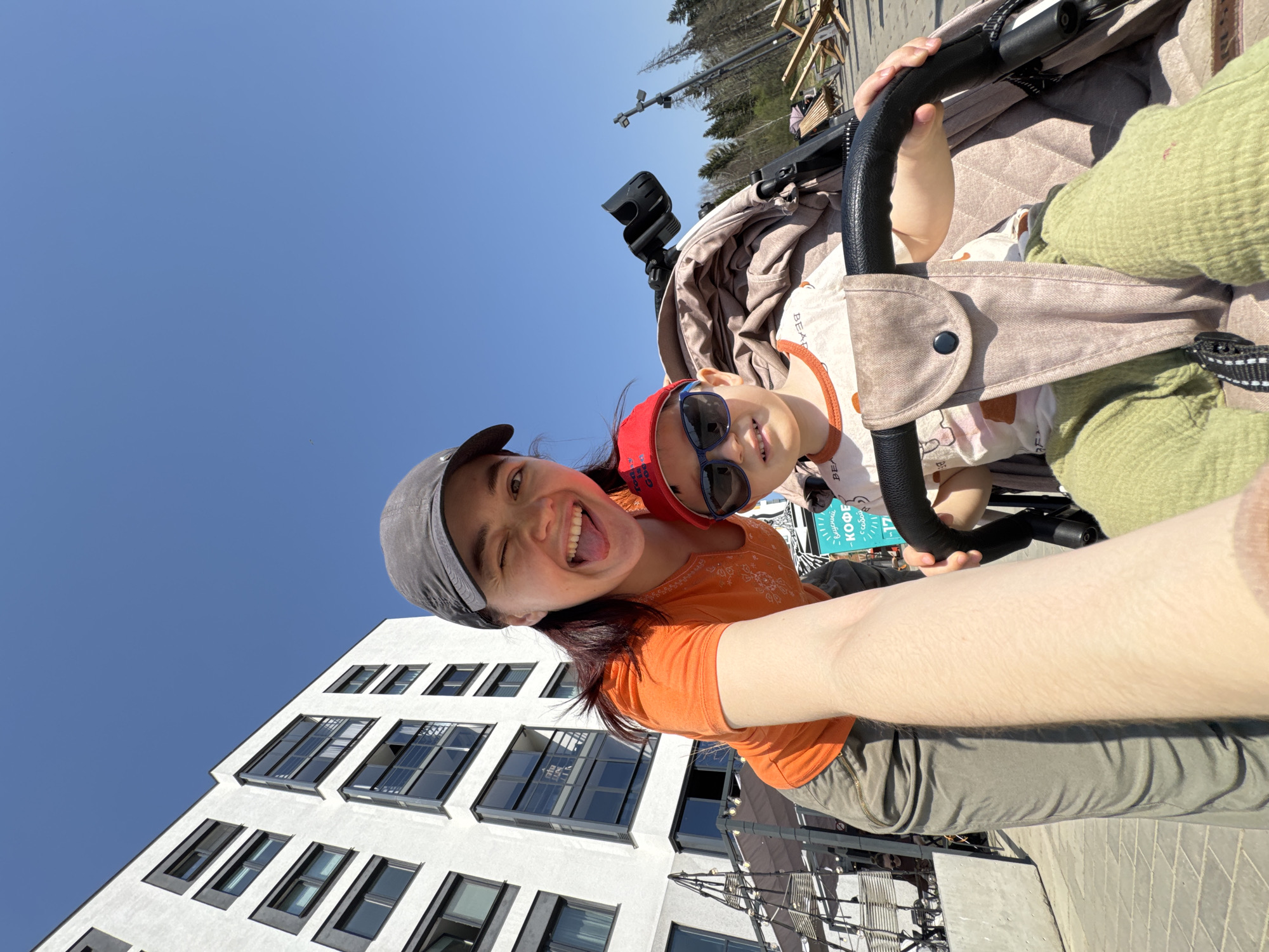 A person with long brown hair, wearing an orange t-shirt, grey pants, and a grey cap, is taking a selfie with a young child in a stroller. The child is wearing sunglasses and a red cap. They are outdoors, with a modern white building featuring dark windows visible in the background. Based on the Cyrillic signage and architectural style, the location appears to be in Izhevsk, Russia. The sky is a clear blue. The person is holding the camera at arm's length, capturing themselves and the child in the stroller against the backdrop of the city.