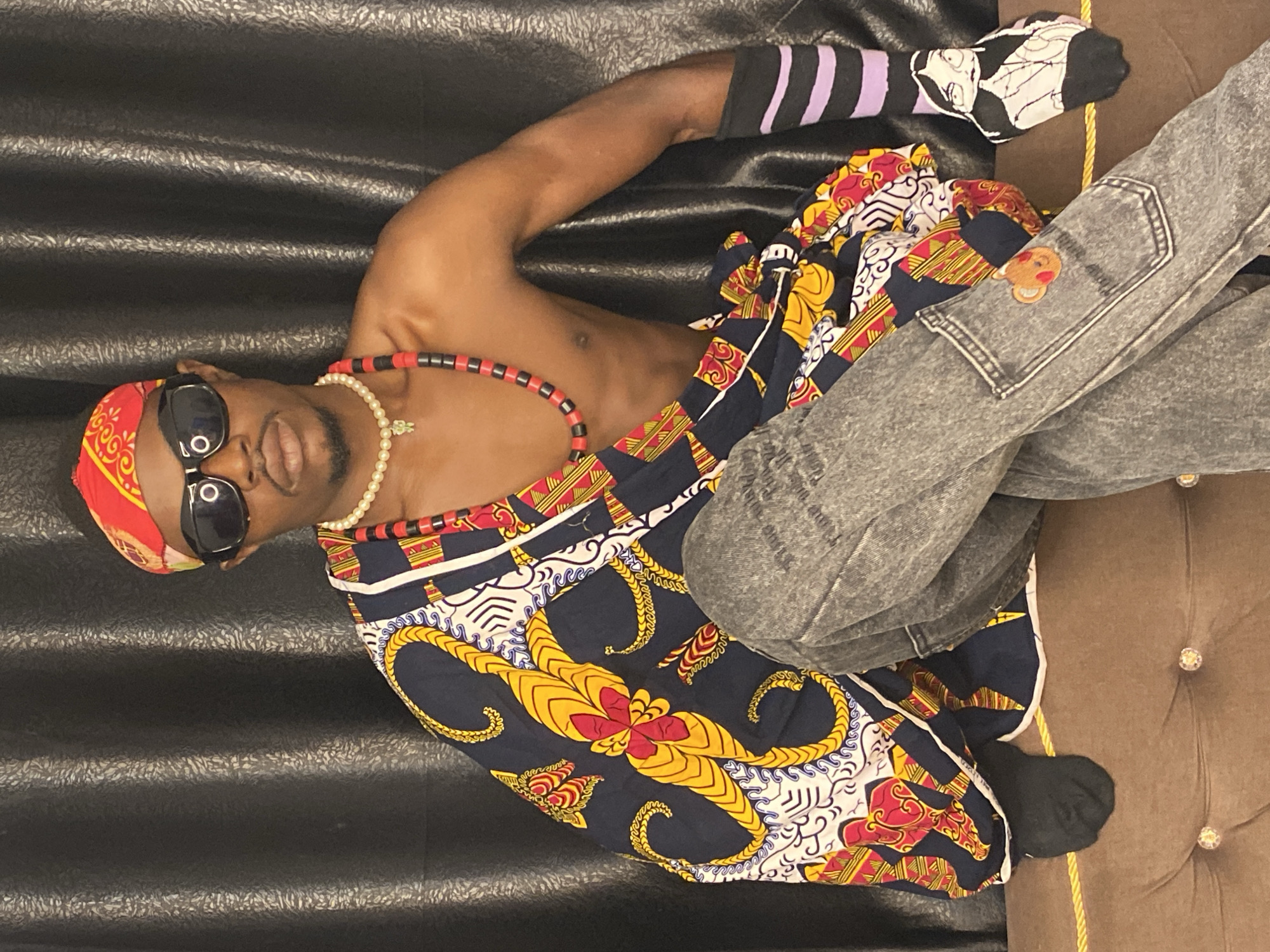 A person with dark skin, wearing a red and yellow bandana, sunglasses, a pearl necklace, and a beaded necklace, is seated against a black curtain. They are wearing a colorful fabric draped over their upper body, grey denim jeans with a bear patch on the pocket, black socks, and striped socks with a doll image on the foot. The person is leaning on a brown cushioned surface with decorative buttons. There is no visible indication of the city of Abakaliki, Nigeria, or any other specific location.