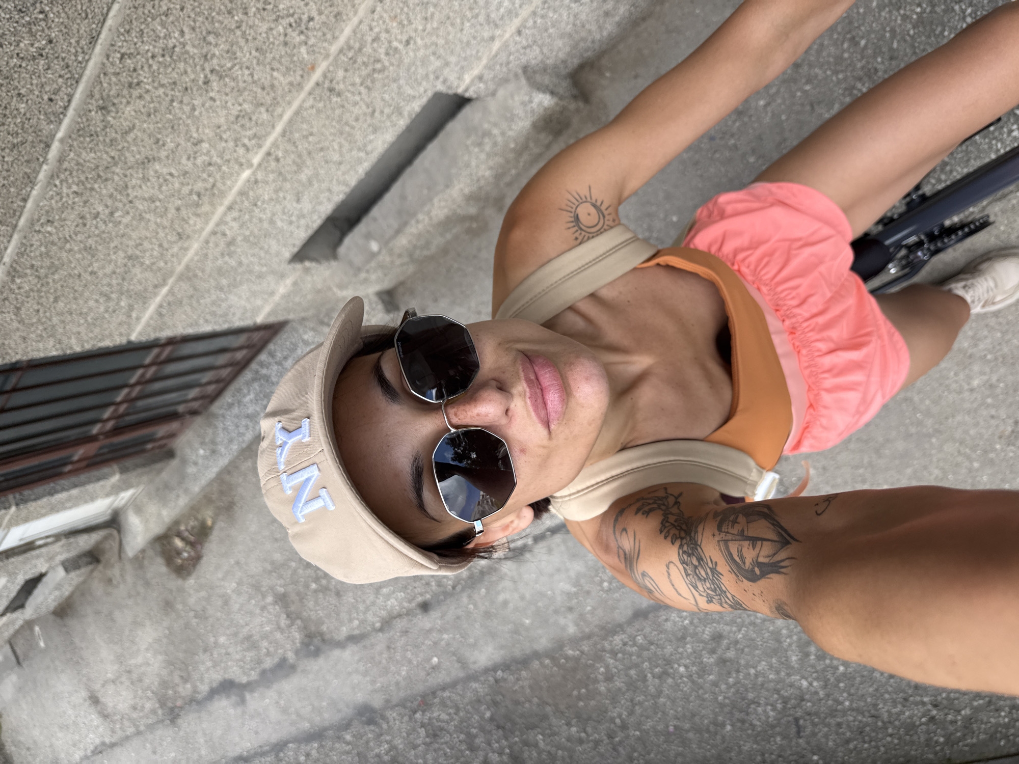 The image shows a woman riding a bicycle on a street in the city of Pleven, Bulgaria. She is wearing a beige baseball cap with "NY" embroidered on it, sunglasses, a light beige tank top with an orange trim, and coral-colored shorts. The woman has tattoos on her left arm, including a sun and moon design on her shoulder and a detailed image of two faces on her upper arm. She is looking at the camera with a slight smile. The background shows a stone building with barred windows and a paved street. The perspective suggests the photo was taken from a high angle, possibly by the woman herself.