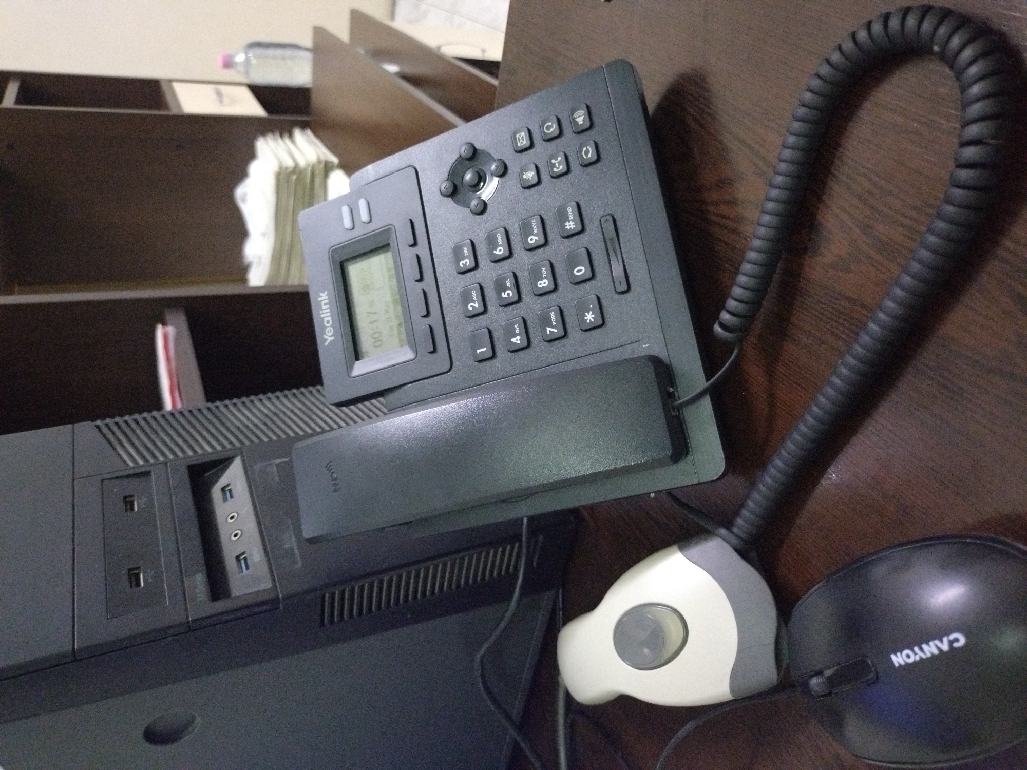 The image depicts a dark brown wooden desk surface covered with office equipment. Centered is a black Yealink office phone with a handset on its cradle. The phone's display reads "Yealink" and "00:17 Tue 20 May." A black coiled phone cord extends from the phone. Adjacent to the phone are parts of a black computer case on the left and a two-toned beige and black computer mouse (Canyon) on the right. The desk surface is dark and the lighting is subdued. A bookshelf with papers and a water bottle is visible in the background. There is no information available to determine if this image was taken in Stara Zagora, Bulgaria.