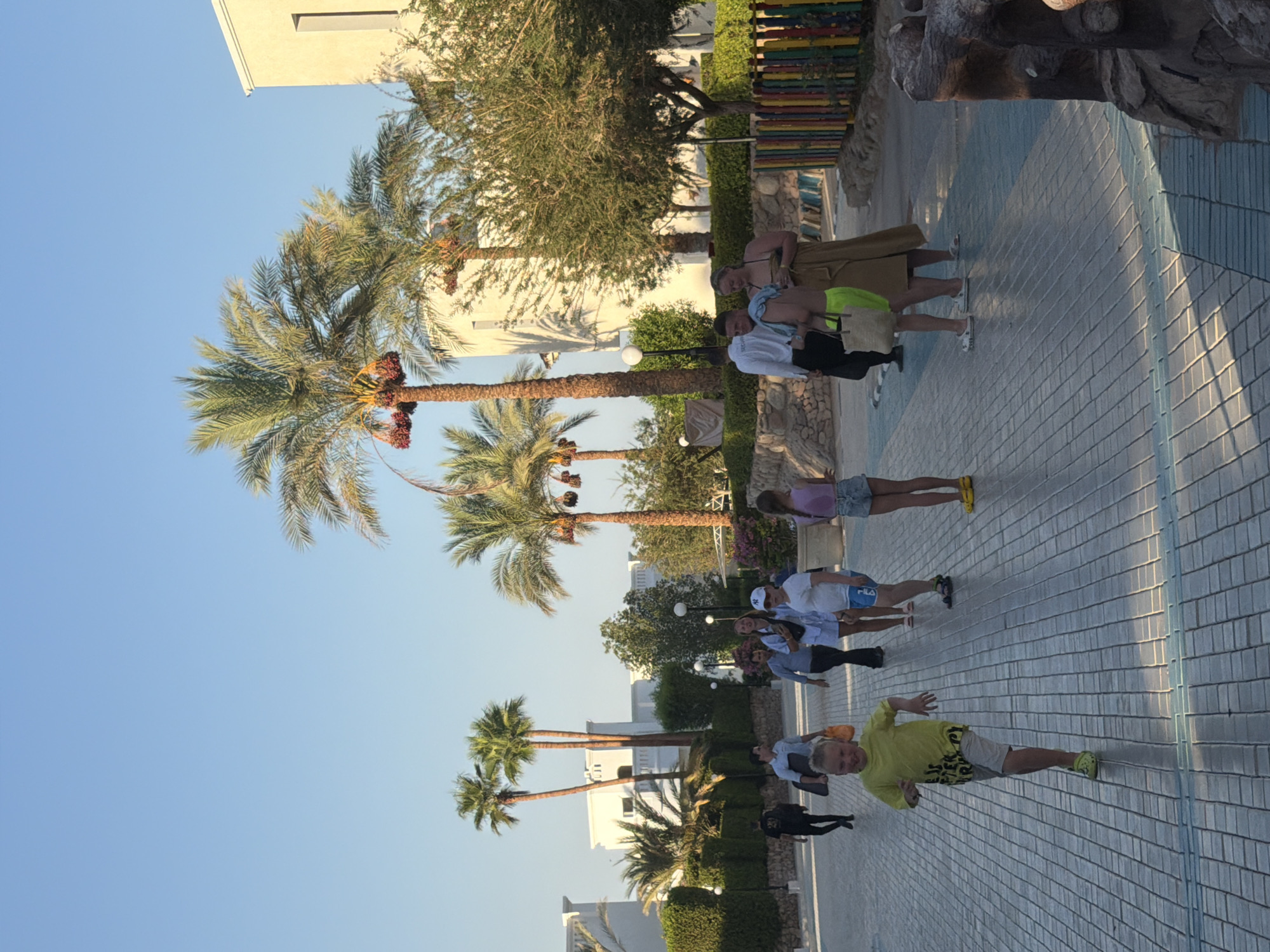 The image depicts a sunny daytime scene in Sharm El Sheikh, Egypt, likely within a resort or hotel complex. The foreground features a paved walkway with a blue and gray tiled pattern. A young boy in a yellow t-shirt and light-colored shorts is running towards the viewer, smiling broadly. Several other people are walking in the background, including adults and children.
The environment is characterized by lush palm trees with dates hanging from them, and manicured hedges. In the background, several white buildings with distinct architectural features are visible, suggesting a resort setting. The sky is clear and blue. To the right, a colorful wooden fence separates the walkway from a rocky area, possibly an entrance to a themed attraction or a decorative element. The lighting indicates it is either early morning or late afternoon, with long shadows cast on the ground. The overall atmosphere is relaxed and suggests a vacation or leisure setting.