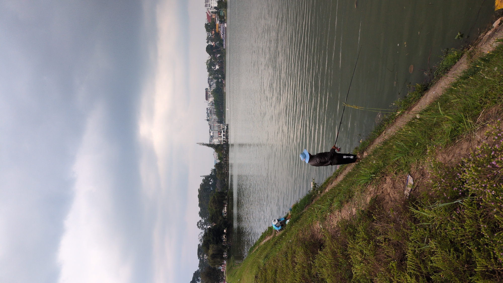 A man fishes along the shoreline of a lake. He is wearing a blue hat and dark clothing. A fishing rod is extended over the water. Other buildings and trees are visible across the lake. The sky is cloudy. The photograph was taken at Xuan Huong Lake in Da Lat, Vietnam. Another person sits along the edge of the lake further down the shoreline. A concrete lip is visible above the water line. Grass and other vegetation grow on the shore.