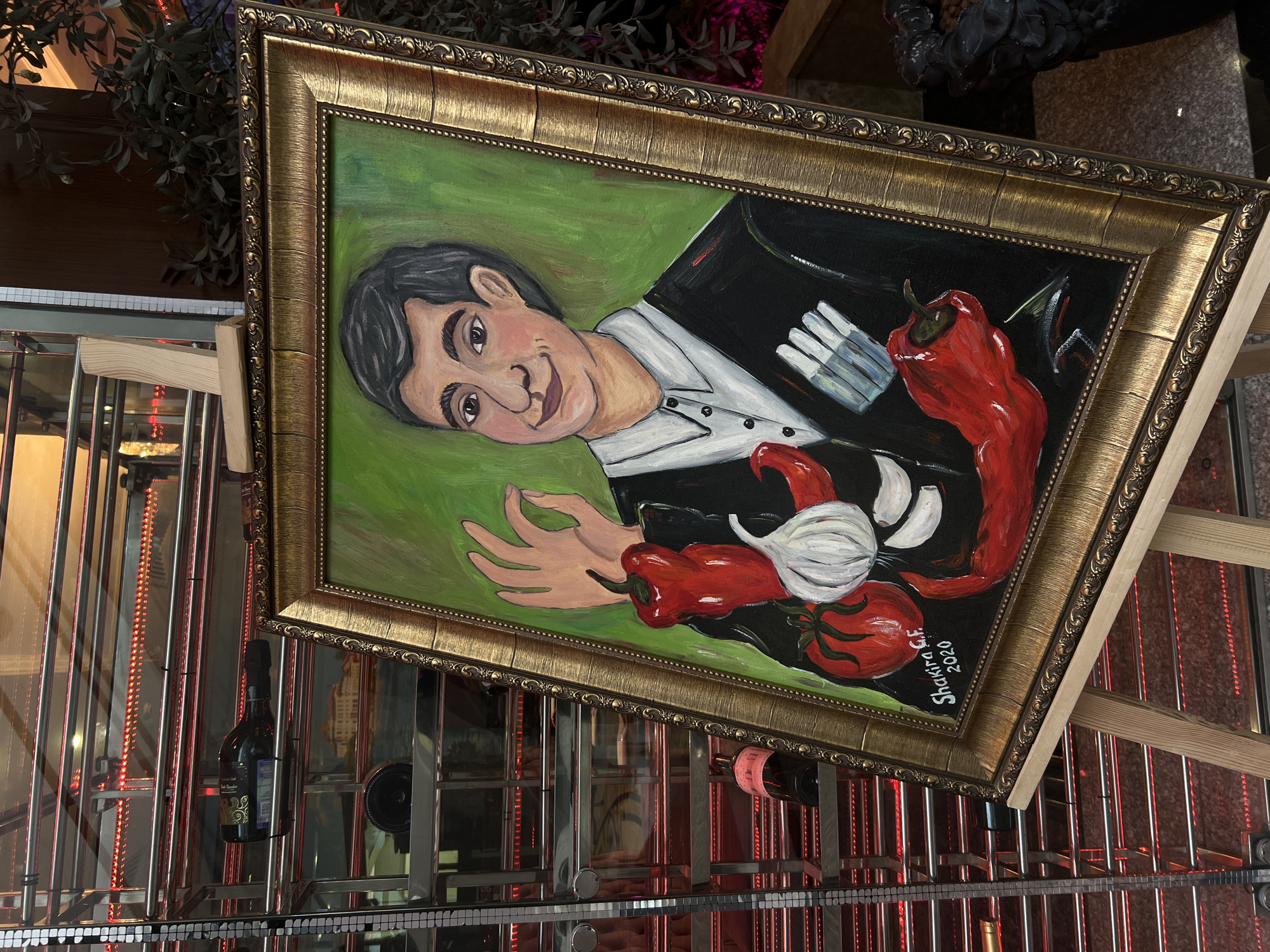 The image shows a framed painting of a man in a suit accompanied by vegetables, peppers, and garlic. The man is light-skinned with dark hair and wearing a white collared shirt with a black jacket. A disembodied hand is on the left side. The background is green and the painting has a gold-colored ornate frame. The signature on the artwork reads "Shakira G.F. 2020". The painting is placed on an easel on a rack. The rack has lighting and there are wine bottles also stored on the rack. The painting is located at a restaurant in Kazan, Russia.