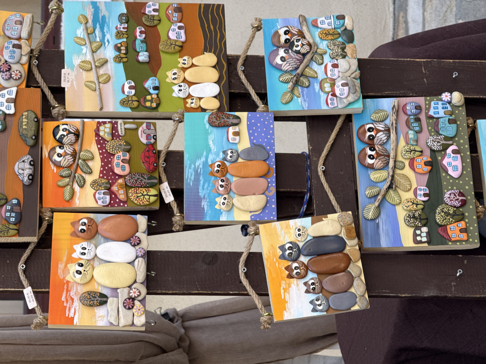 The image shows a wooden stand displaying various handcrafted art pieces made from painted stones. These art pieces depict scenes with stylized animals like cats and owls, as well as miniature houses and trees. The stand is set up outdoors, likely at a market or craft fair, with a dark fabric draped around its base. One of the art pieces has a price tag that reads "25,00€ 48,89 лв.", indicating prices in Euros and Bulgarian Lev, suggesting the location is in Bulgaria. The overall aesthetic is colorful and whimsical. The scene is likely located in Bansko, Bulgaria, a popular tourist destination known for its crafts and festivals.