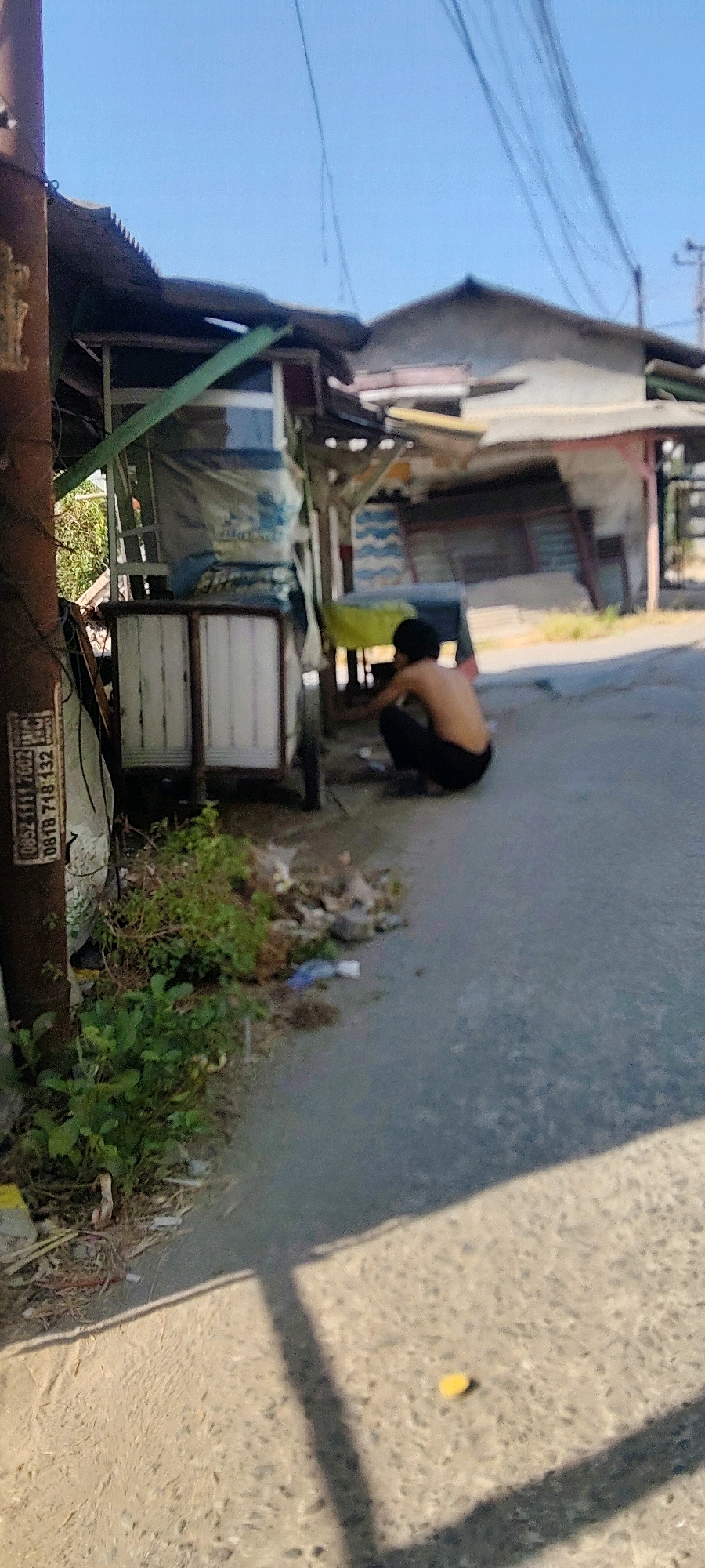 A shirtless person wearing black pants is crouched on the ground next to a mobile cart. The cart appears to be made of wood and metal, with some fabric coverings. The person is engaged in an activity that is not clearly discernible from the image. The cart is positioned on an asphalt road, with a dirt shoulder to the left covered in weeds and debris. In the background, there are several dilapidated structures, possibly stalls or makeshift buildings, with corrugated metal roofs. The sky is clear and blue. The scene is set outdoors, likely in a street or market area in Mekarmukti, Indonesia, given the presence of the cart and the style of the buildings. The overall environment appears to be somewhat neglected, with visible trash and unkempt vegetation.