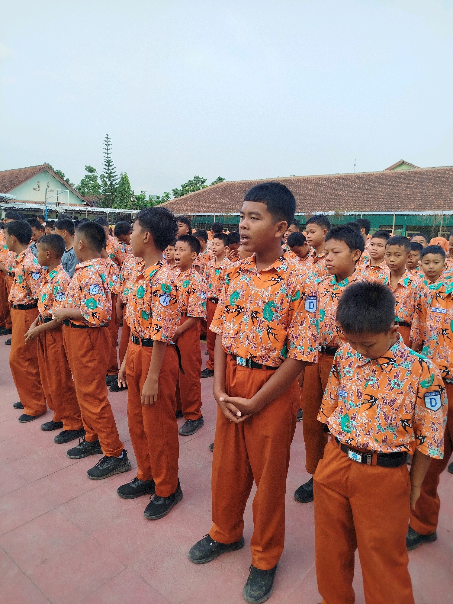 In Karangsambung, Indonesia, a group of young male students in matching orange uniforms with a bird and floral pattern stand in formation outdoors. The uniforms consist of short-sleeved button-up shirts with patches and solid orange pants, paired with black belts and black shoes. The students, primarily of Indonesian ethnicity, appear to be participating in a school assembly or activity on a paved surface in front of low-rise school buildings with tiled roofs. The sky is overcast.