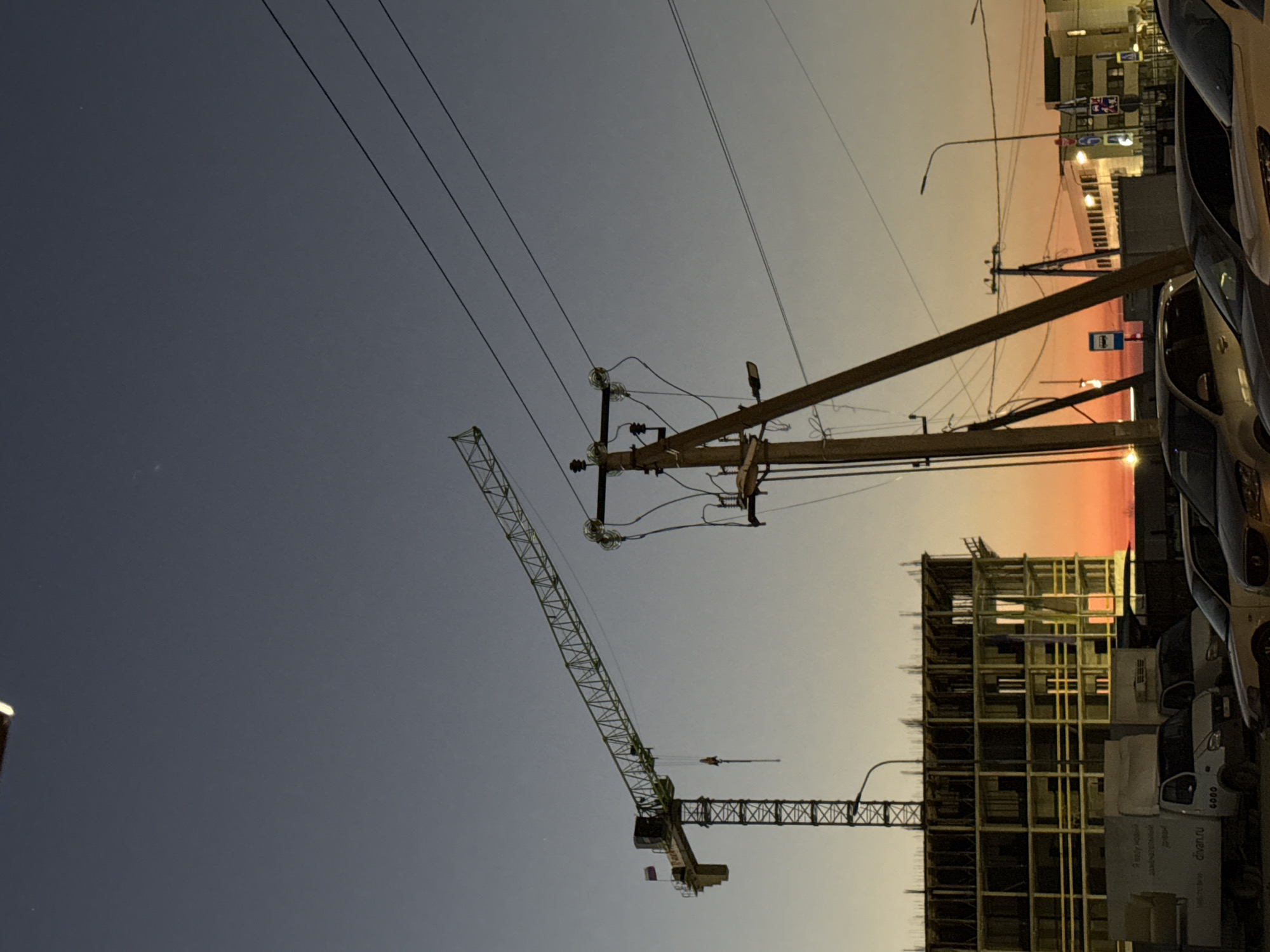 A low-angle, color photograph depicts a cityscape at dusk, featuring a construction site with a crane. The sky transitions from a deep blue-gray in the lower left to a warm orange-pink at the upper right horizon. In the foreground, electrical wires stretch across the sky, connected to a utility pole with insulators and other attachments. A construction crane is situated towards the left of the frame, its lattice structure extending upwards. To the right of the crane, a building under construction is visible, its framework of beams and supports partially completed. Parked cars are arranged along the right edge of the image, obscuring some of the street and buildings behind them. The scene suggests urban development against a backdrop of changing daylight.