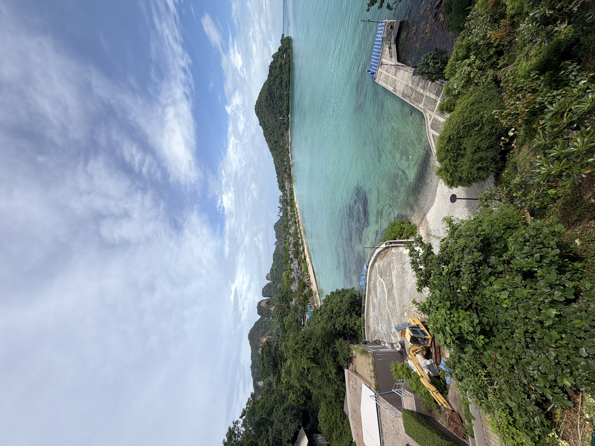 This is a high-angle, wide-shot photograph of a scenic coastal view in Ao Nang, Thailand. The foreground features lush green foliage and a construction area with a yellow excavator. A concrete circular platform is visible, with a building to the left. A paved walkway leads to a concrete pier extending into the turquoise ocean. In the middle ground, a crescent-shaped sandy beach is lined with resort bungalows and palm trees, backed by a dense green forest and dramatic, rocky limestone karsts that are characteristic of the region. A larger, tree-covered island juts out into the ocean on the right side of the frame. The sky is partly cloudy with scattered white clouds against a blue backdrop. The overall scene depicts a tropical coastal landscape with elements of development and natural beauty.