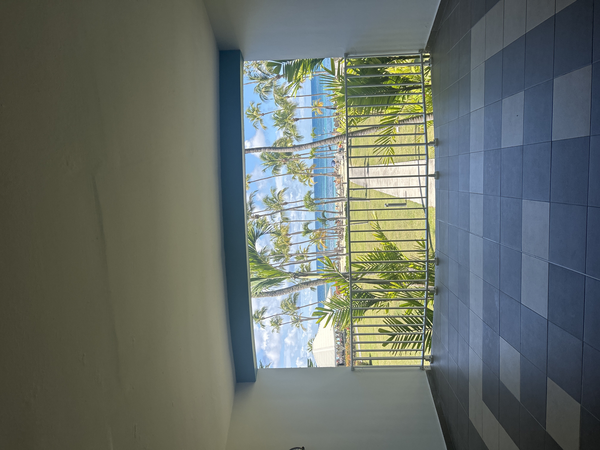 From an interior perspective, a balcony view showcases a tropical beach scene. A series of palm trees lines the beach, where people are visible. The balcony features a metal railing, with a tiled wall to the right. A glimpse of a building is seen through the foliage. The image captures a tranquil atmosphere. While Sainte-Anne has tropical views, there is insufficient information to precisely locate it.