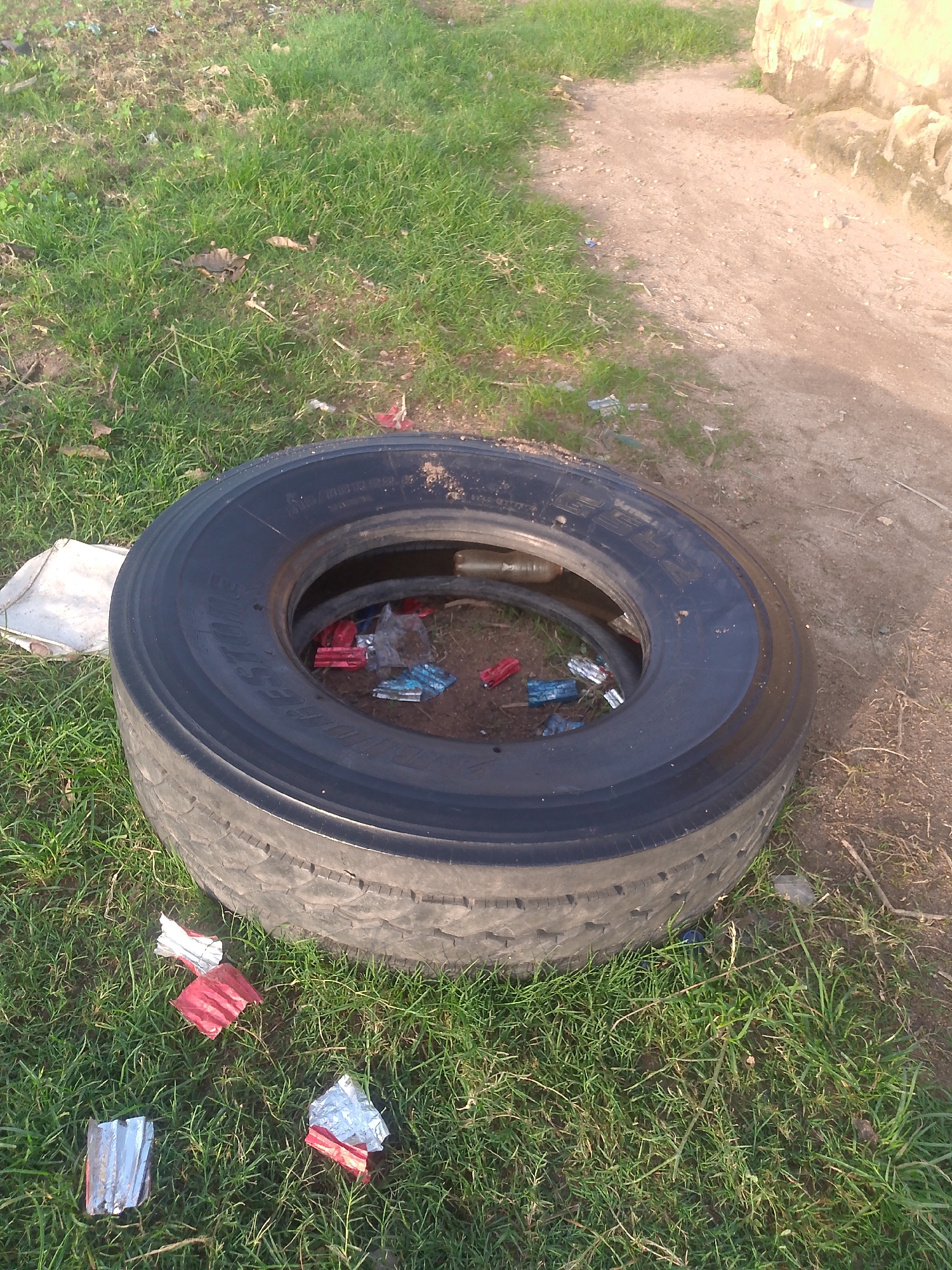 A large, black truck tire lies on its side in a grassy area next to a dirt path. The tire is positioned in a way that its opening faces upwards. Inside the tire, several small, colorful objects are visible, appearing to be remnants of candy wrappers or small plastic items, in shades of red and blue. A white sack or bag is partially visible behind the tire to the left. The ground is covered in green grass, with patches of dry earth and scattered twigs. A dirt path is visible to the right of the tire, leading away from the viewer, with a low, light-colored concrete structure at the edge of the path. The overall scene suggests an outdoor, possibly informal, setting in the city of Jalingo, Nigeria, characterized by the presence of discarded items.