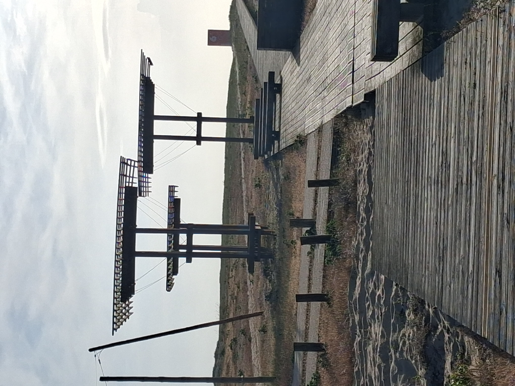 Here is a description of the image:
The image depicts a landscape scene with several notable elements. In the left portion, there is a series of dark, rectangular structures arranged in a vertical stack. These structures are held together with wire. The background of these structures is an overcast sky.
To the right, a wooden boardwalk stretches into the distance, composed of planks. Adjacent to the boardwalk, a grassy area extends outwards, and a set of wooden posts run across the landscape. There is a long dark horizontal line that appears to traverse the composition from the lower left to the upper right, intersecting the wooden posts and structures.
The scene appears to be in Figueira da Foz, Portugal.