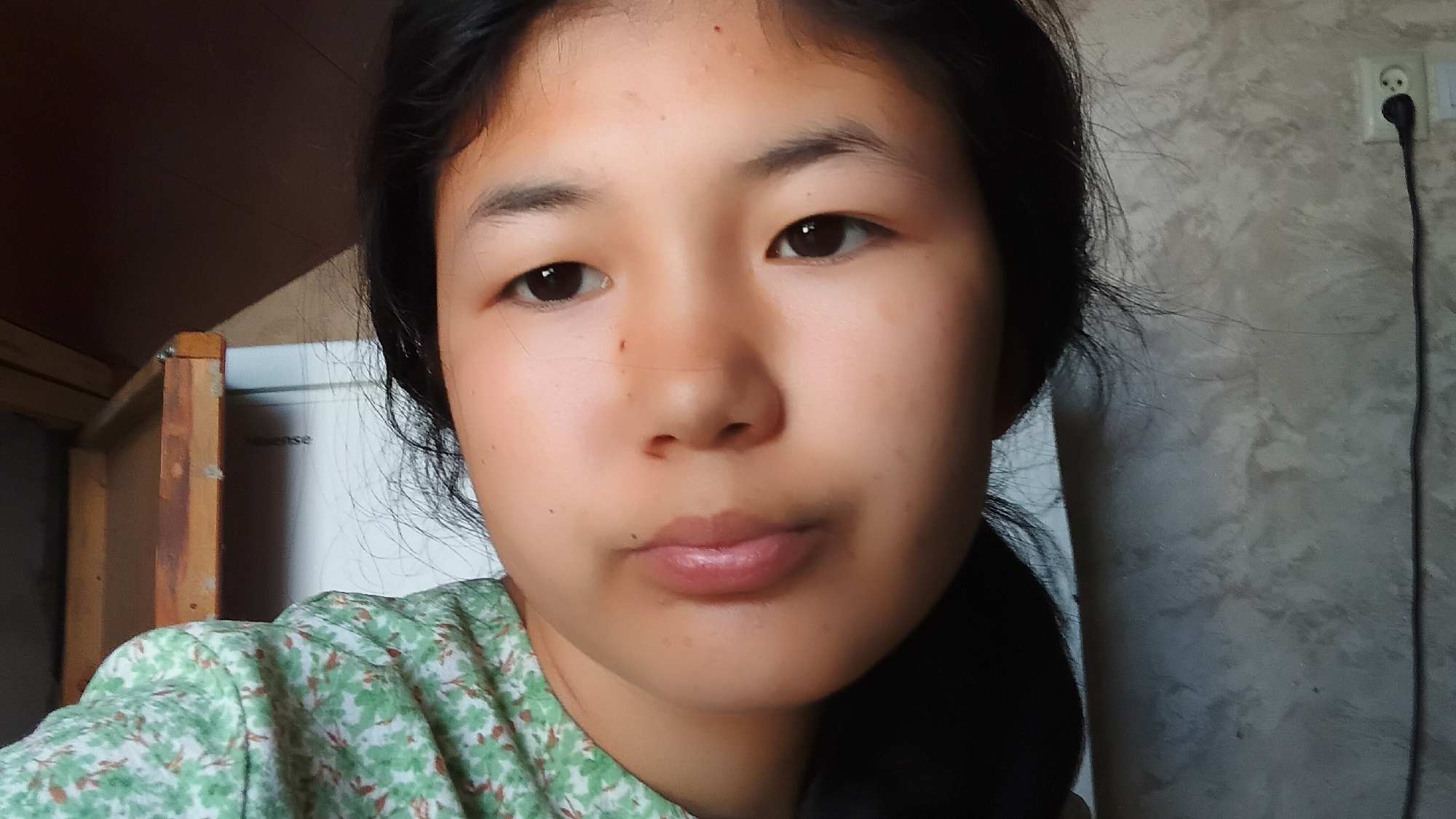 In an indoor setting, a young East Asian female with dark hair is positioned centrally, looking directly at the camera. She is wearing a green floral print shirt. Her face is illuminated by soft lighting. Behind her to the left, a white refrigerator with the brand name "Hisense" is visible, partially obscured by a wooden frame structure. To her right, a grey textured wall is present, featuring a white electrical outlet with a black cord extending downwards. The scene appears to be in a residential space in the city of Uchkun, Kyrgyzstan.
