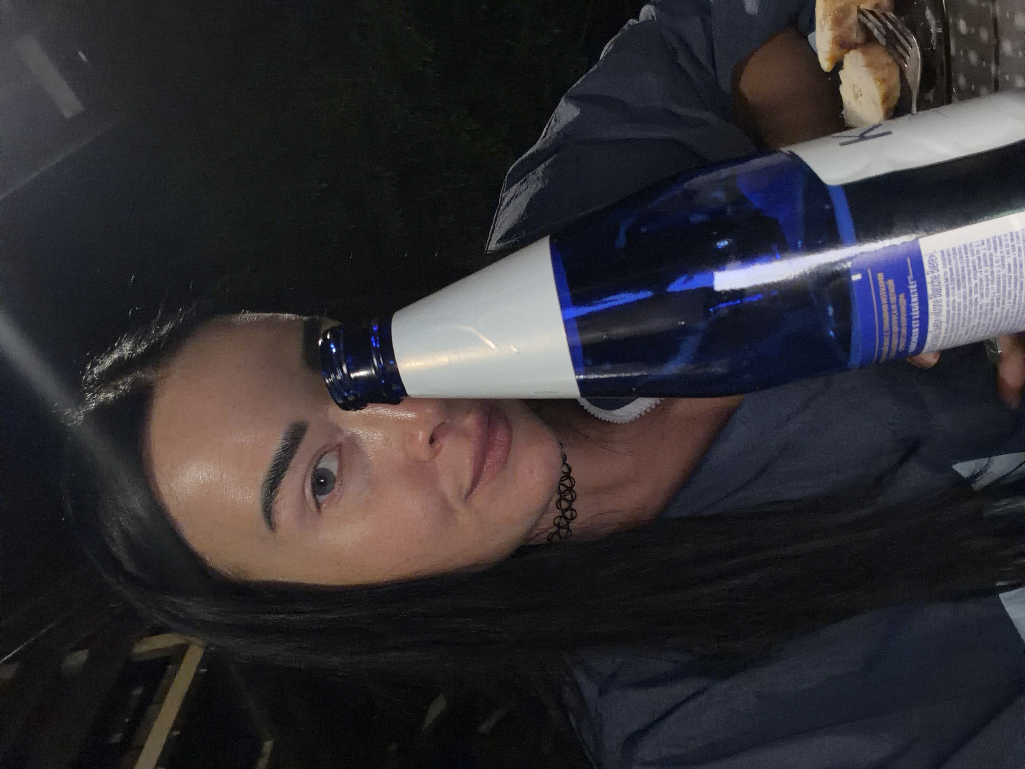 A woman with dark hair and a choker necklace is holding a blue glass bottle in front of her face, obscuring her right eye. She is wearing a dark blue shirt and is seated at a table where bread and a fork are visible. The background is dark and appears to be outdoors, possibly at night. The location is not identifiable as Sochi, Russia.