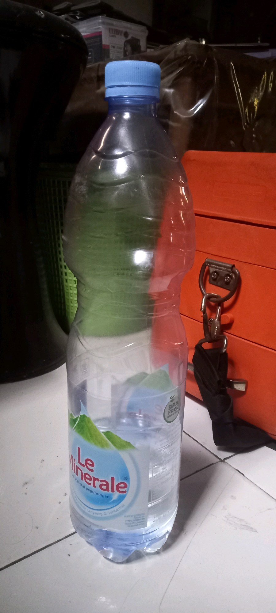 A plastic bottle of Le Minerale bottled water stands on a tiled floor. The bottle is mostly full, with a blue cap. In the background, there is an orange toolbox with a metal latch and a black strap attached to it. To the left of the bottle, a dark, blurry object, possibly furniture, is visible. The scene appears to be indoors, in an area with a utilitarian feel, like a garage or workshop.