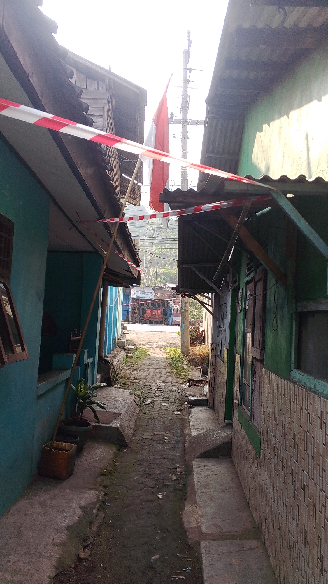 The image depicts a narrow alleyway in Bandar Lampung, Indonesia, likely in a residential or commercial district. Red and white striped tape is stretched across the alley, possibly indicating a closed-off area or a celebration. A flagpole with the Indonesian flag (red and white) is visible, suggesting a national holiday or event. The alley is paved with interlocking bricks and is lined with modest, weathered buildings on either side. One building on the left is painted blue and appears to be a dwelling with windows and a porch. The building on the right is painted green and also shows signs of habitation, with tiled walls and windows. The environment is somewhat cluttered, with visible debris and plants growing along the sides of the alley. In the distance, the alley opens up to a street with a red vehicle and a sign. The overall impression is of a lived-in, somewhat aged urban environment.