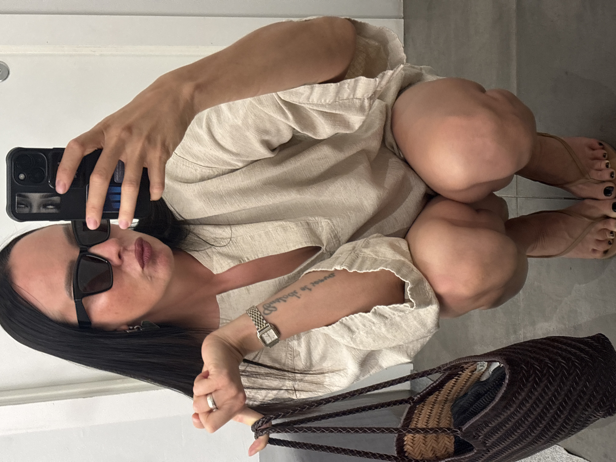 A woman is taking a selfie, squatting on a gray tile floor. She wears black sunglasses, a light-colored linen shirt, and a silver wristwatch on her left wrist, which also has a tattoo. Her toenails are painted black. She holds a black smartphone in her right hand. A brown woven bag is in front of her. The location is indicated to be Chaikovsky, Russia, based on the question. A white door is visible in the background on the left.