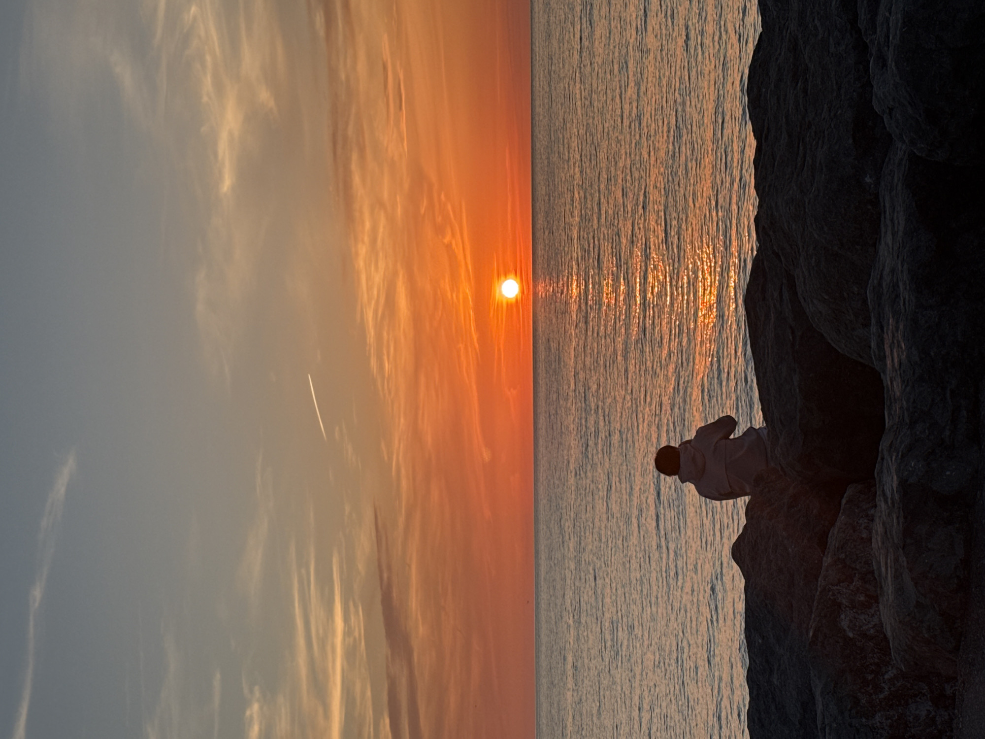The image captures a breathtaking coastal sunset in Batumi, Georgia. The sun, a vivid orange sphere, is low on the horizon, dominating the center of the frame and casting a warm, radiant glow across the sky and water. The sky, primarily on the left, transitions from soft blues and grays to vibrant oranges and reds near the sun, adorned with wispy, elongated clouds catching the golden light. A subtle contrail is visible high in the sky.
The right half of the image is dominated by the serene waters of the sea or ocean, reflecting the sun's brilliance in a shimmering path of light that extends from the horizon towards the foreground. On the far right, large, dark rocks form a dramatic silhouette against the bright water and sky. Perched upon these rocks, a lone individual is visible in silhouette, seated and seemingly observing the mesmerizing sunset. The person is wearing a light-colored garment. The scene conveys a sense of tranquility and awe, highlighting the beauty of nature at dusk. There is no visible text in the image.