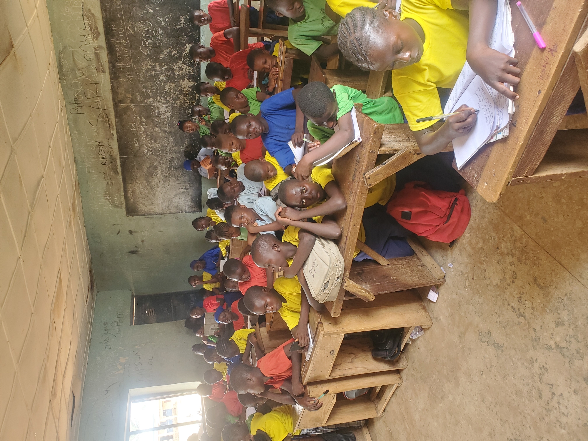 A classroom in Akwanga, Nigeria, is filled with students dressed in yellow, green, red, and blue shirts sitting at wooden desks. They are writing in notebooks with pens. In the background, graffiti covers the walls, including names like "RAPSON" and "GREDOO." A red backpack sits on the floor near a desk. The flooring has a rough, worn appearance. Sunlight streams in through a window.