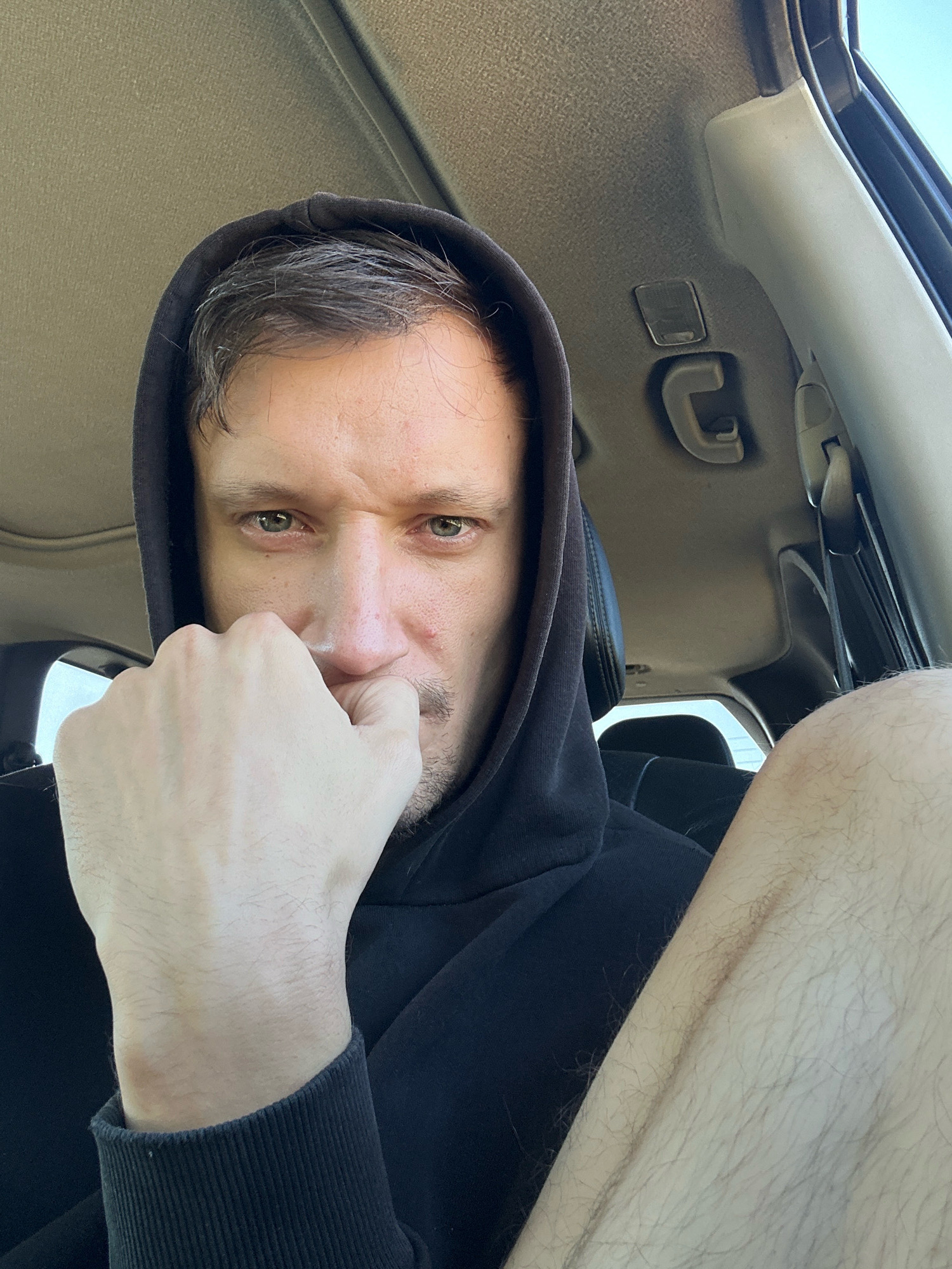 In this close-up, self-taken photograph, a man with short, dark hair and light green eyes is positioned inside a car. He is wearing a black hooded sweatshirt, and the hood is pulled up, framing his face. His left hand is raised, with his fist clenched, and his knuckles are pressed against his mouth and nose, obscuring part of his face. His gaze is directed towards the camera with an intense expression. The interior of the car is visible in the background, including the car's ceiling, a grab handle, and part of a seat. The lighting suggests it is daytime, with sunlight possibly coming from the side. The location within the city of Makiv, Ukraine, is not discernible from the visual information provided in the image.