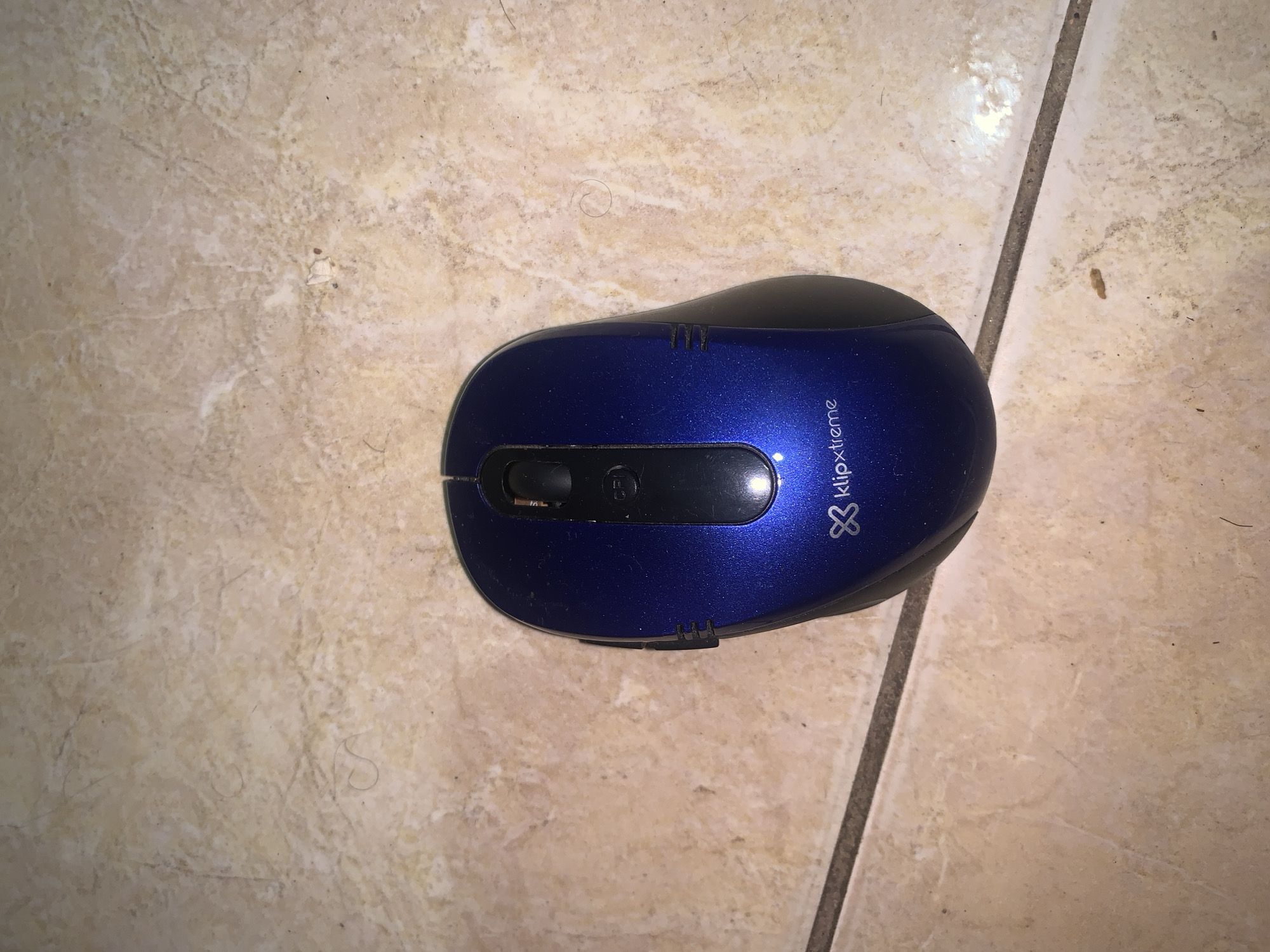 A blue and black Klip Xtreme computer mouse rests on a tiled floor in Santa Ana, Venezuela. The mouse is positioned centrally, viewed from a top-down angle, slightly askew. Its upper shell is a metallic blue, transitioning to matte black on the sides and front where the scroll wheel and buttons are located. The "klipxtreme" brand name and its distinctive "8"-like logo are visible in white text on the right side of the mouse. A black scroll wheel and a smaller central button are present between the main left and right click areas, which are also black.
The floor consists of light beige or cream-colored tiles with a subtle, irregular marbled pattern. A dark grout line runs diagonally across the lower right portion of the image, separating two tiles. Scattered across the tiled surface are various small specks of dirt, dust, and strands of hair, indicating a common indoor setting. The lighting appears to be ambient, suggesting an indoor environment without direct harsh sunlight. No human activity or other subjects are visible.