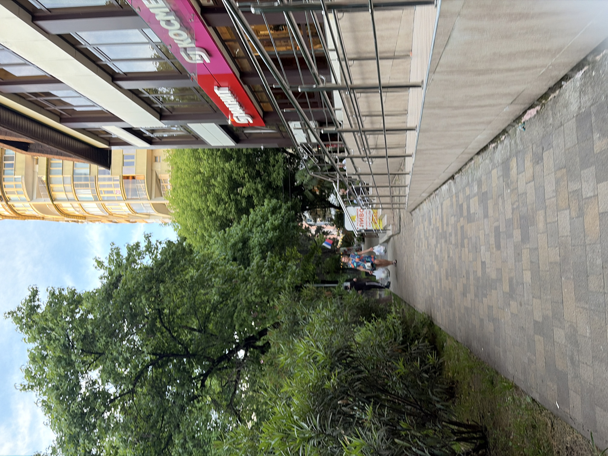 The image depicts a street scene in Sochi, Russia, likely in an urban or commercial area. A paved walkway runs alongside a building with a modern facade, featuring large windows and a prominent pink sign with white Cyrillic lettering that reads "KOCME" (likely part of a larger word like "Kosmetika" or "Kosmetologiya," meaning cosmetics or cosmetology). Another red sign with white Cyrillic lettering, "SHANT," is also visible. To the left of the walkway, lush green trees and bushes create a natural barrier. A woman is walking away from the viewer on the walkway, carrying several white plastic bags. Another person is visible further down the walkway, also carrying bags. In the background, a tall, curved building with balconies and a yellowish exterior is visible, as is a partially visible Russian flag. A ramp with metal railings leads up to the building's entrance. The ground is a brick-patterned pavement. The sky is partly cloudy.