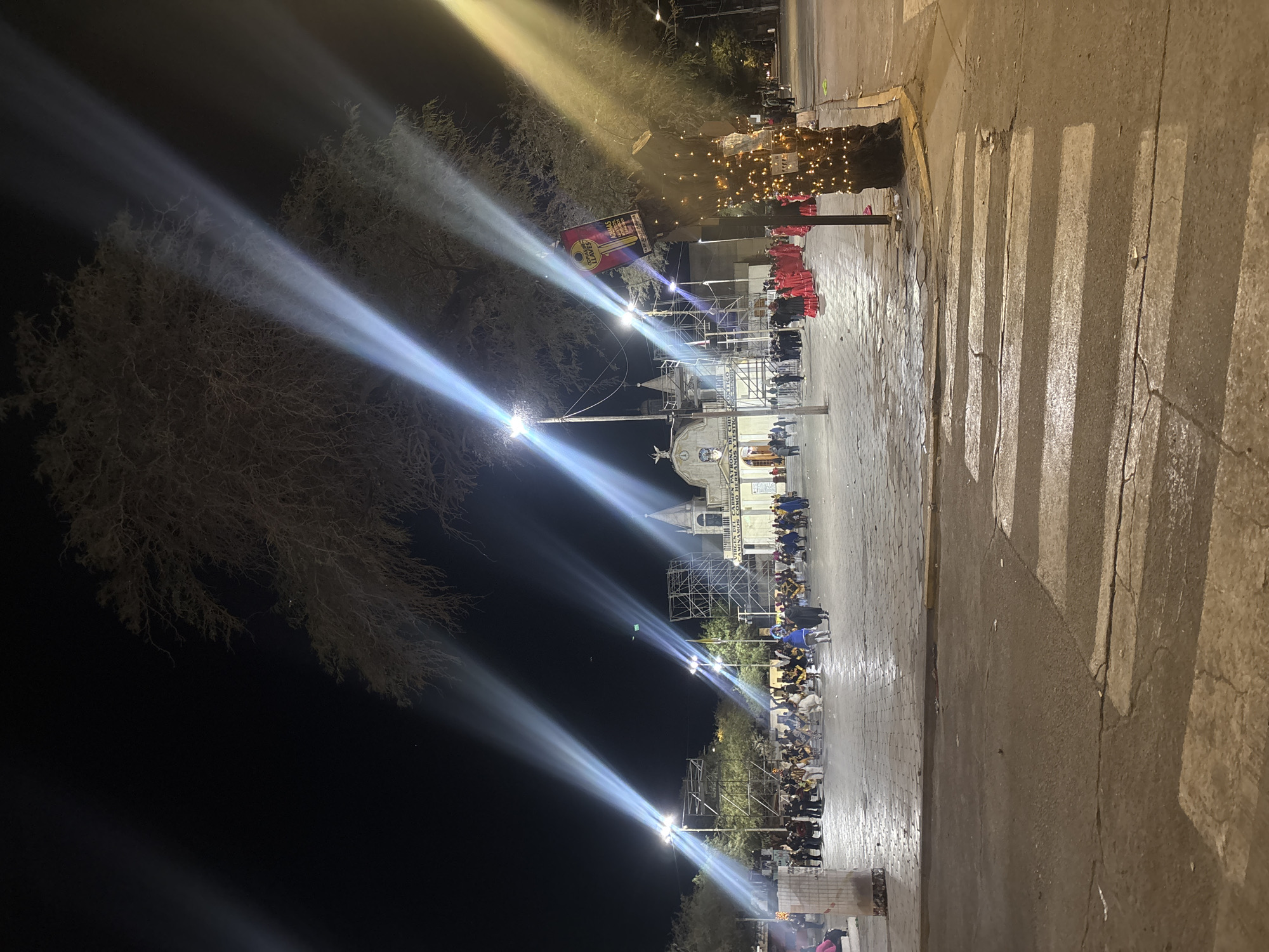 This is a nighttime, long-exposure photograph taken from a street level, looking towards a plaza and a church in Pozo Almonte, Chile. The foreground features a wet asphalt road with a faded white crosswalk. In the mid-ground, a cobblestone plaza is visible, populated by numerous people, some of whom are dressed in colorful costumes, including several individuals in bright red attire. There is a large tree in the foreground with decorative lights wrapped around its trunk, and a banner with text in Spanish hanging from a pole nearby. The background is dominated by a church structure with a steeple and illuminated windows, and scaffolding suggests ongoing construction or setup for an event. Dramatic beams of light, likely from spotlights, cut through the darkness from above, illuminating parts of the plaza and the tree. The overall atmosphere appears to be festive, possibly for a religious or cultural celebration.