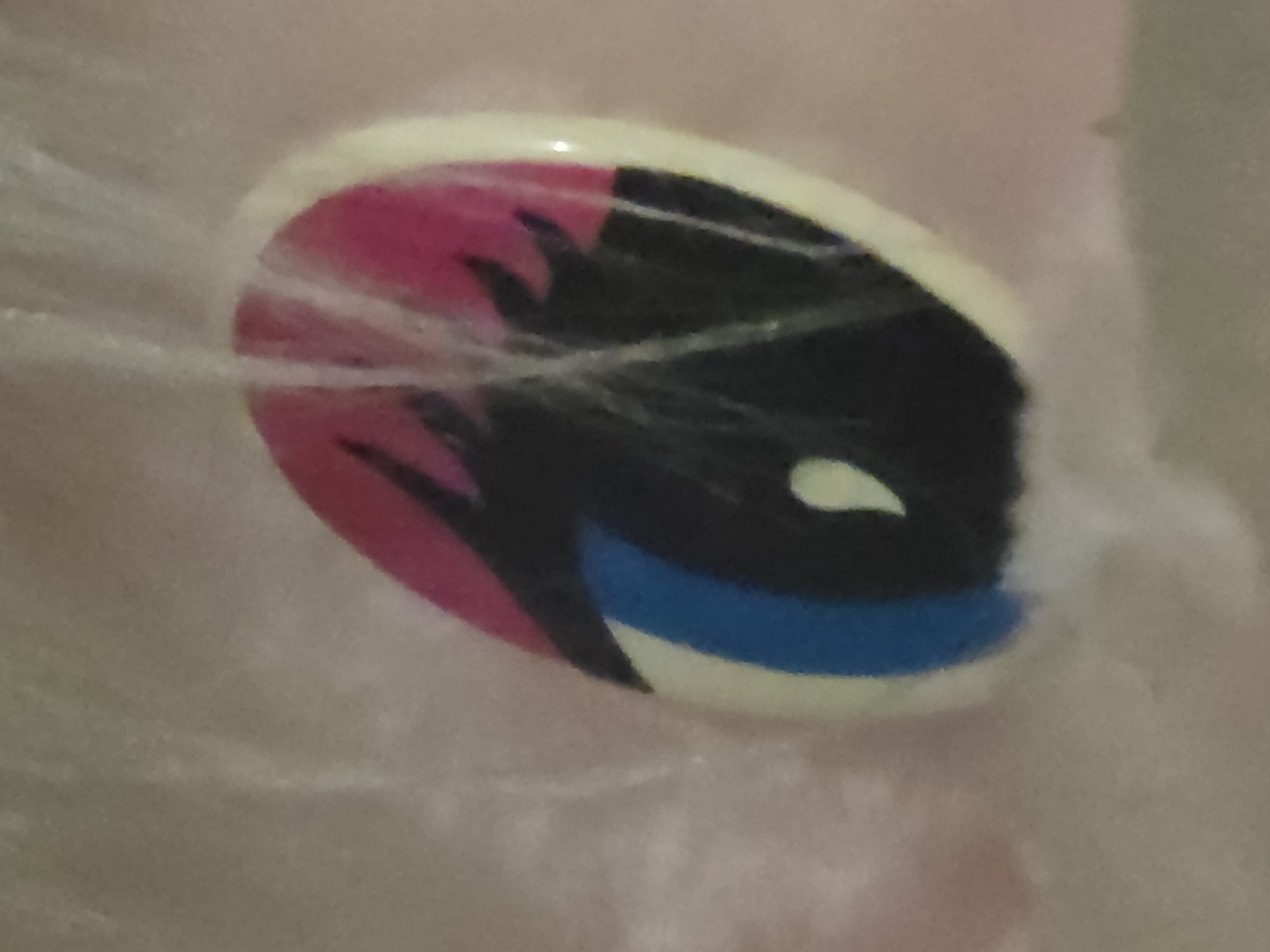 A close-up, angled shot shows a detail of a painted eye. The eye is oval-shaped and outlined in a cream color. The iris is predominantly black, with a bright blue stripe running vertically on the left side. A small, bright yellow teardrop-shaped highlight is positioned in the lower center of the iris. The upper portion of the eye is a vibrant pink, with black, flame-like shapes extending downwards into the iris. Fine, white, wispy fibers are scattered across the surface of the image, partially obscuring the painted eye. The background is a soft, out-of-focus pinkish-tan. The scene is indoors, with no visible environmental details beyond the immediate subject. There are no people present.